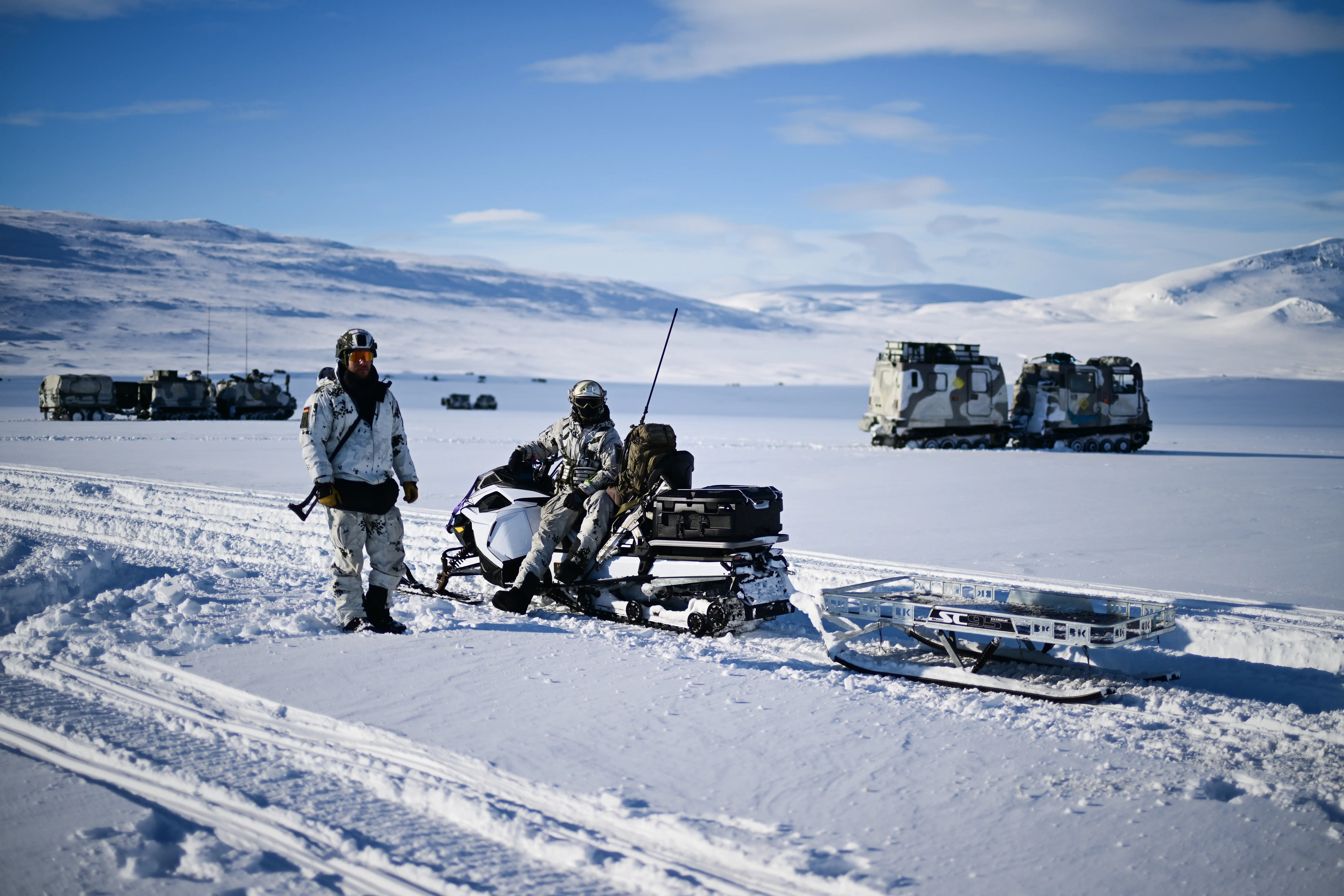 Two figures in camouflage in snow under a blue sky, with one on a snowmobile