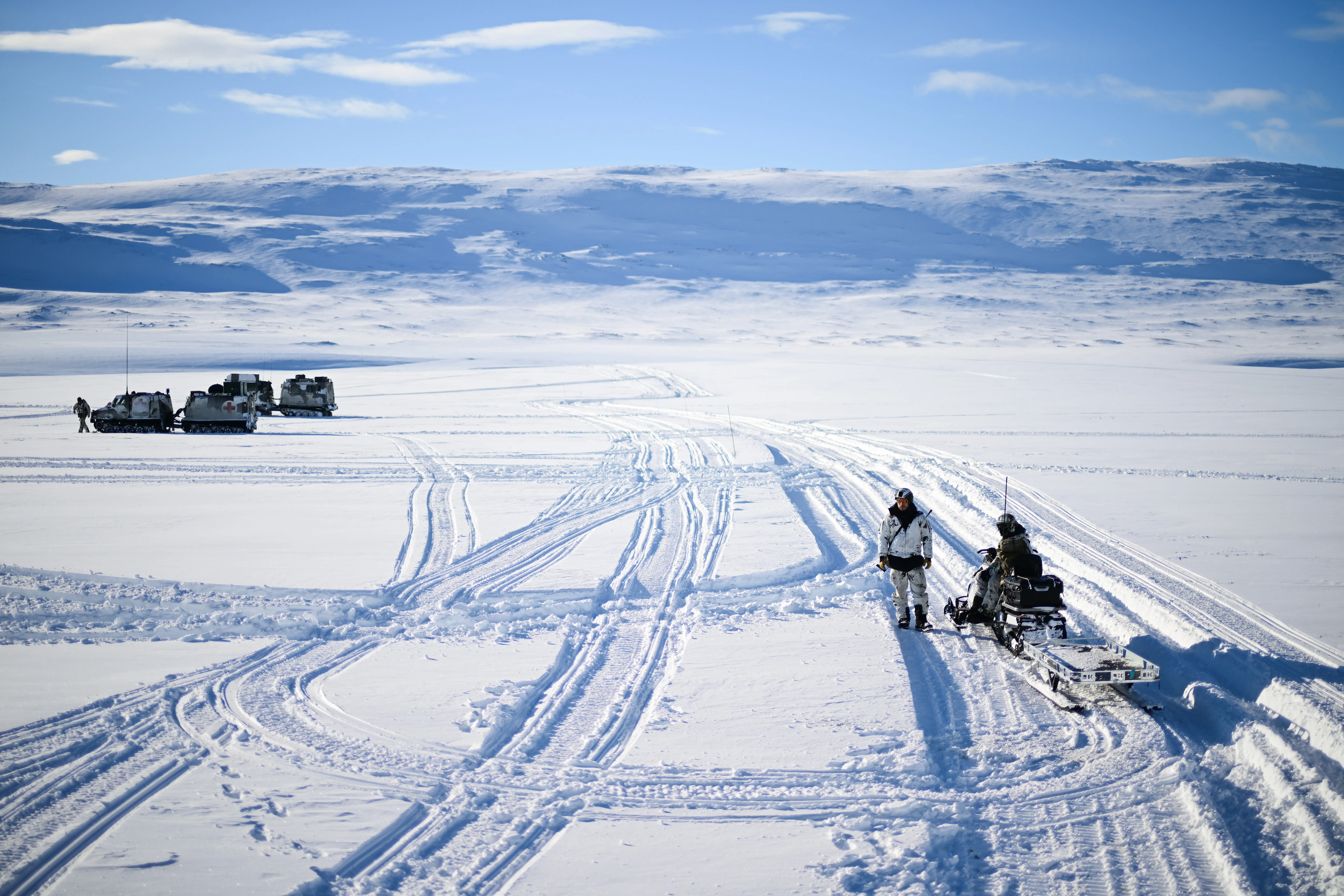 A snowy landscape with humans and a snowmobile in the foreground and vehicles in the distance
