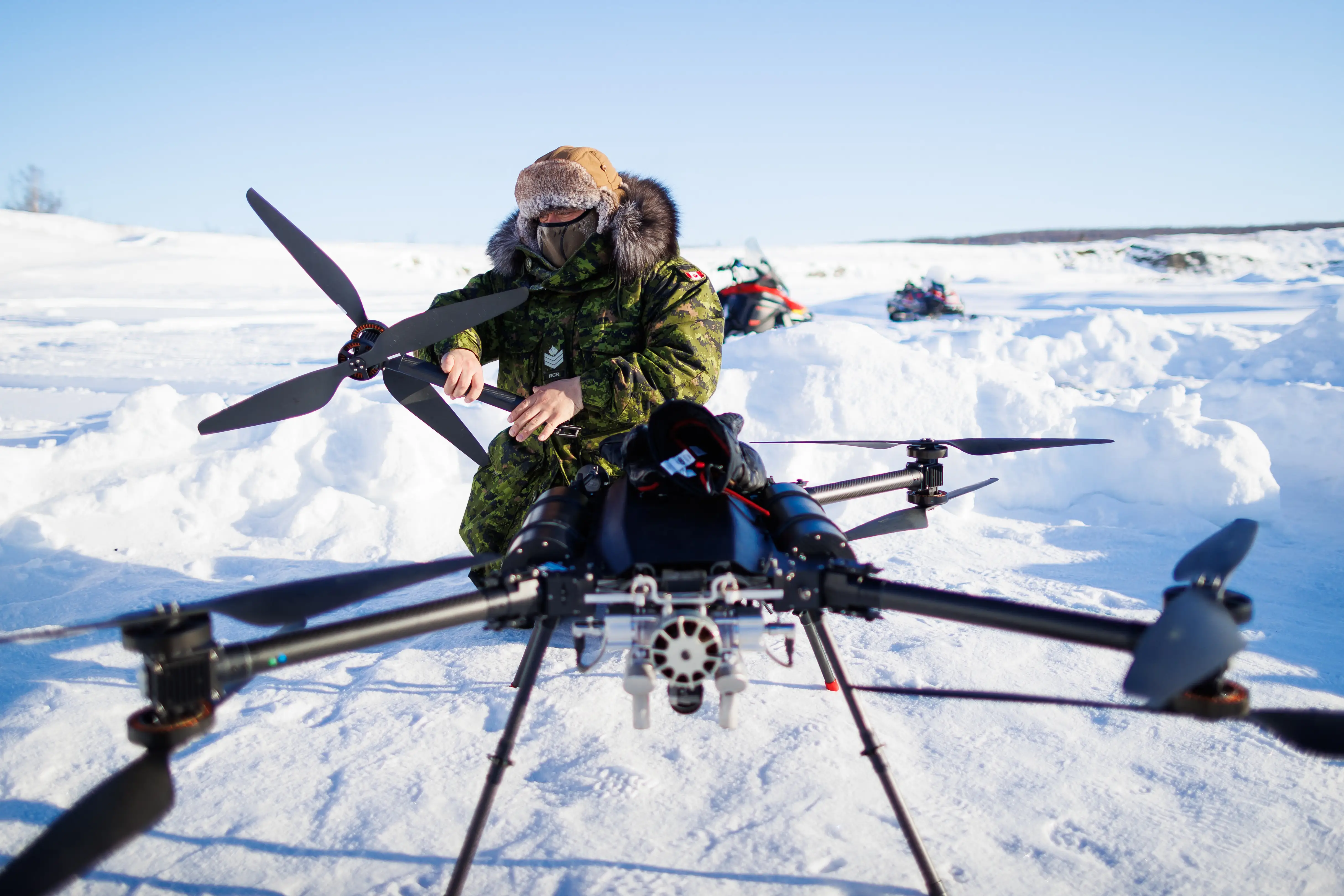 A man in camouflage and a fluffy hat kneels working on a large drone in the snow