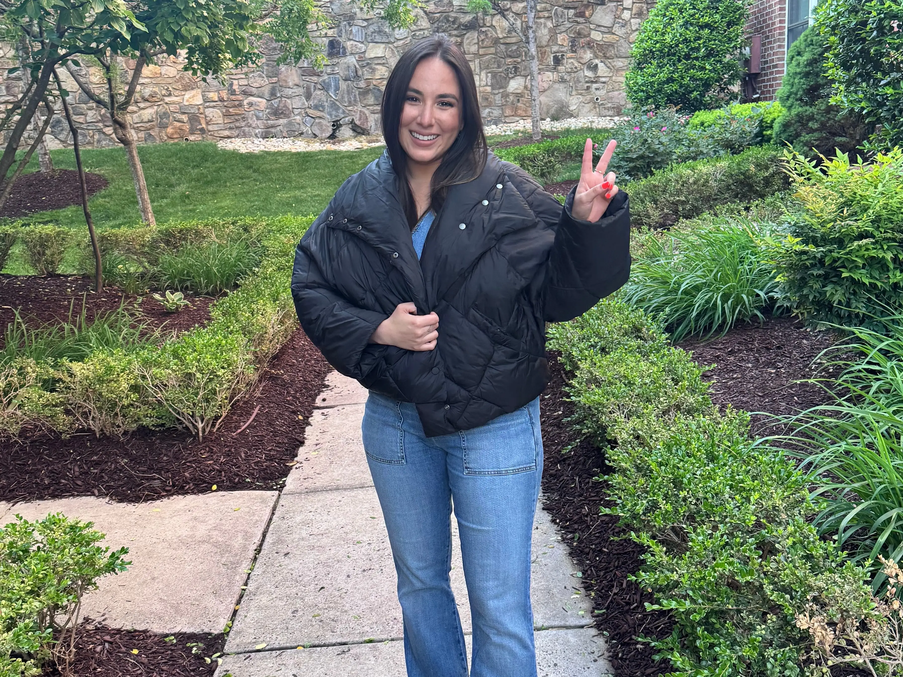 A grinning woman in a blue jumpsuit and a black puff coat holds up a peace sign while standing on a sidewalk.