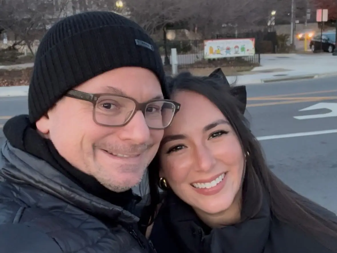 A man wearing glasses and a woman, both dressed in black, smile for a selfie.