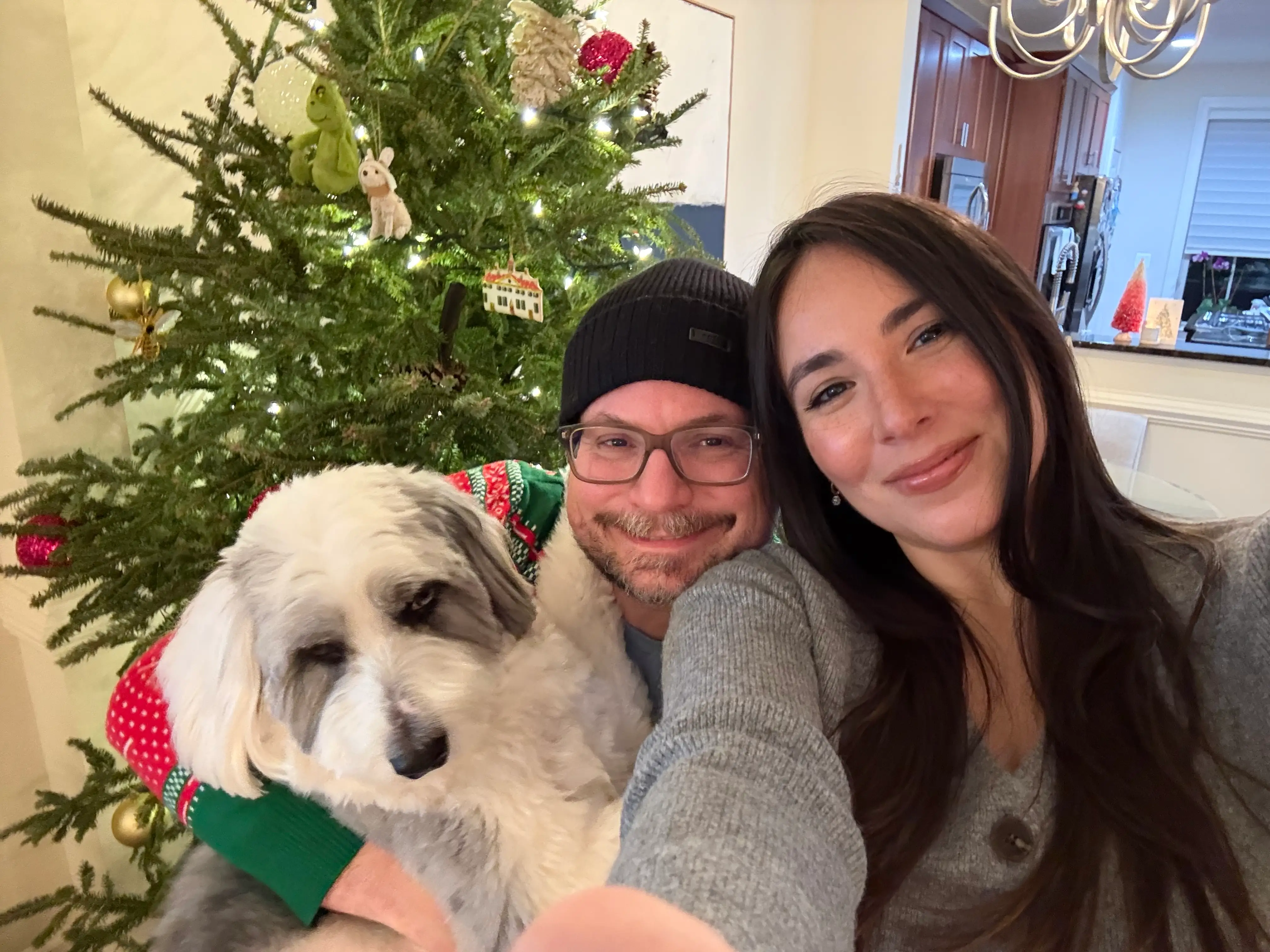 From left to right, a dog, a man, and a woman smile for a selfie in front of a Christmas tree.