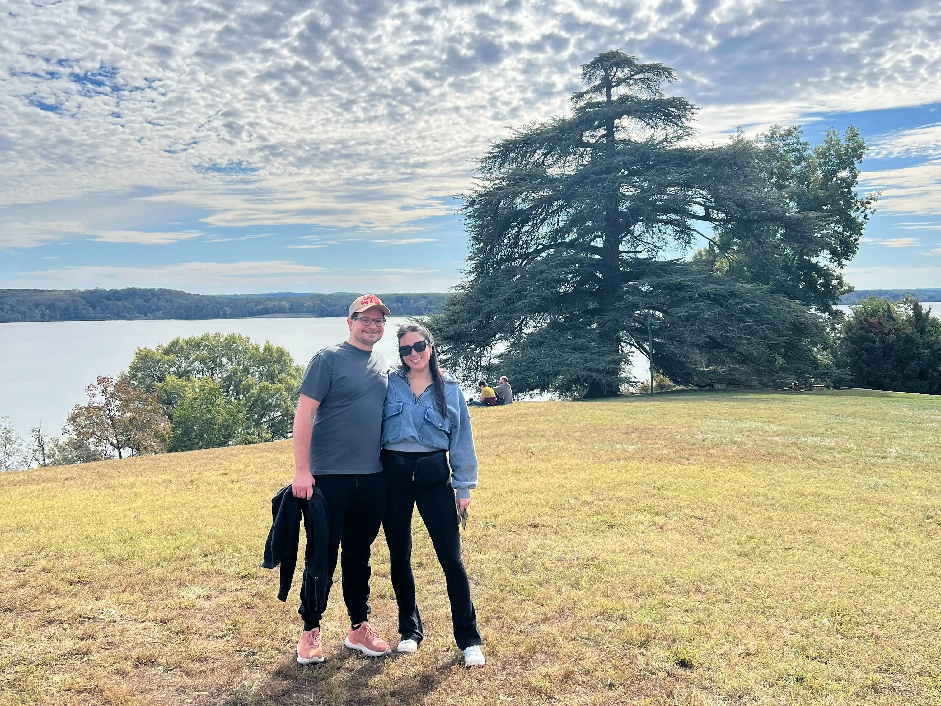 A man and woman pose for a selfie; tall trees stand in the background.