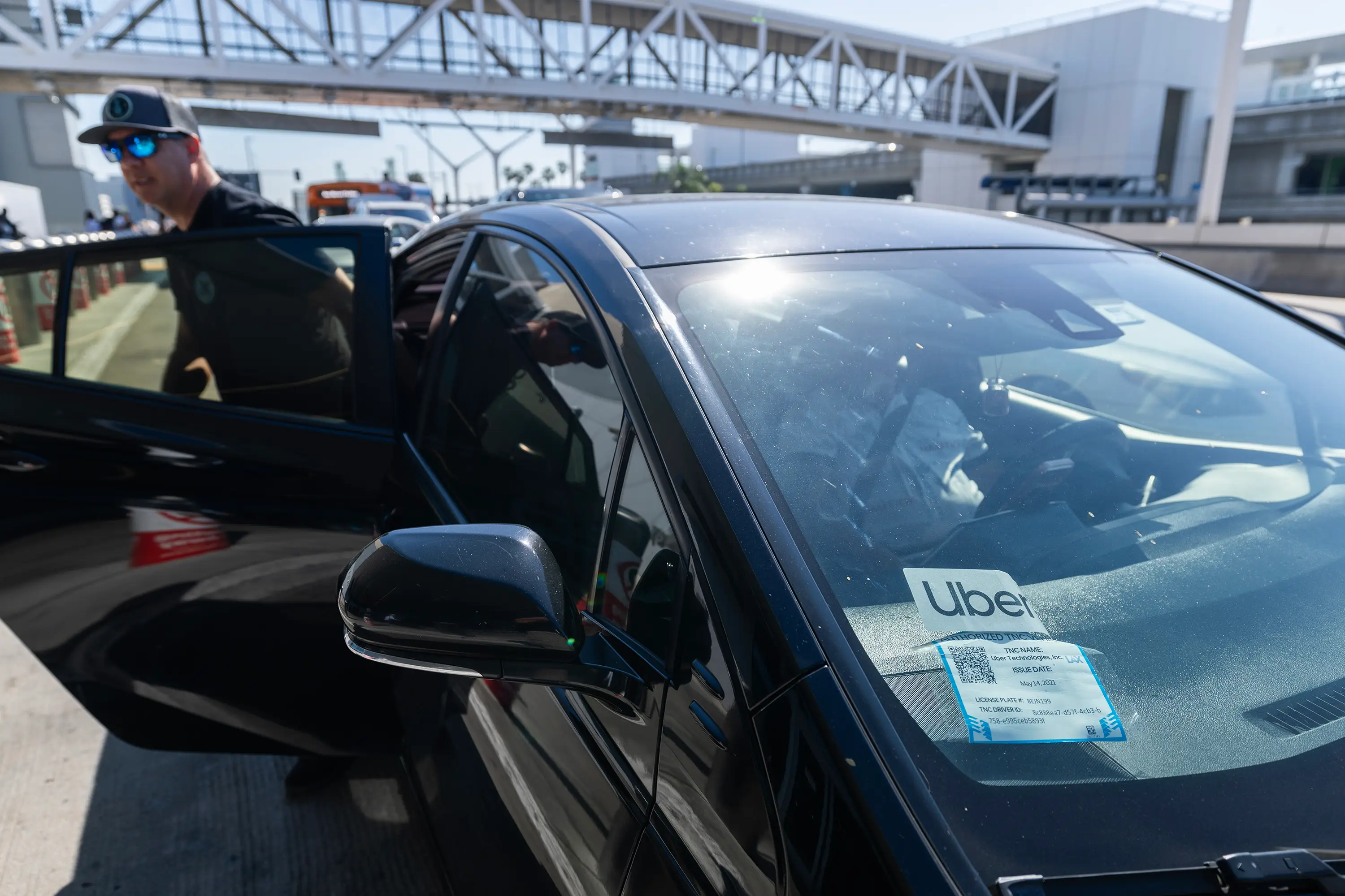 A man exits a compact Uber car while wearing a baseball cap and sunglasses at Los Angeles International Airport.