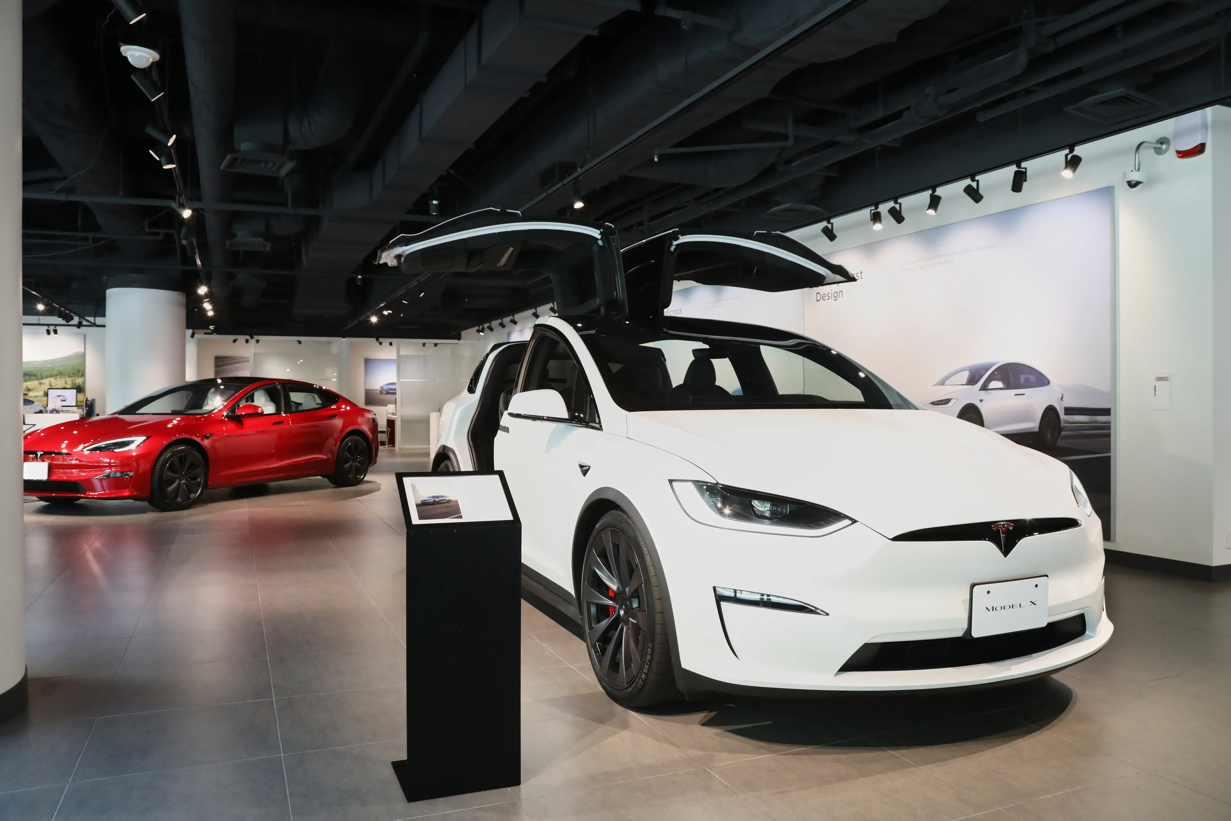 A white Tesla Model X is parked in front of a red Tesla Model S on a showroom floor.