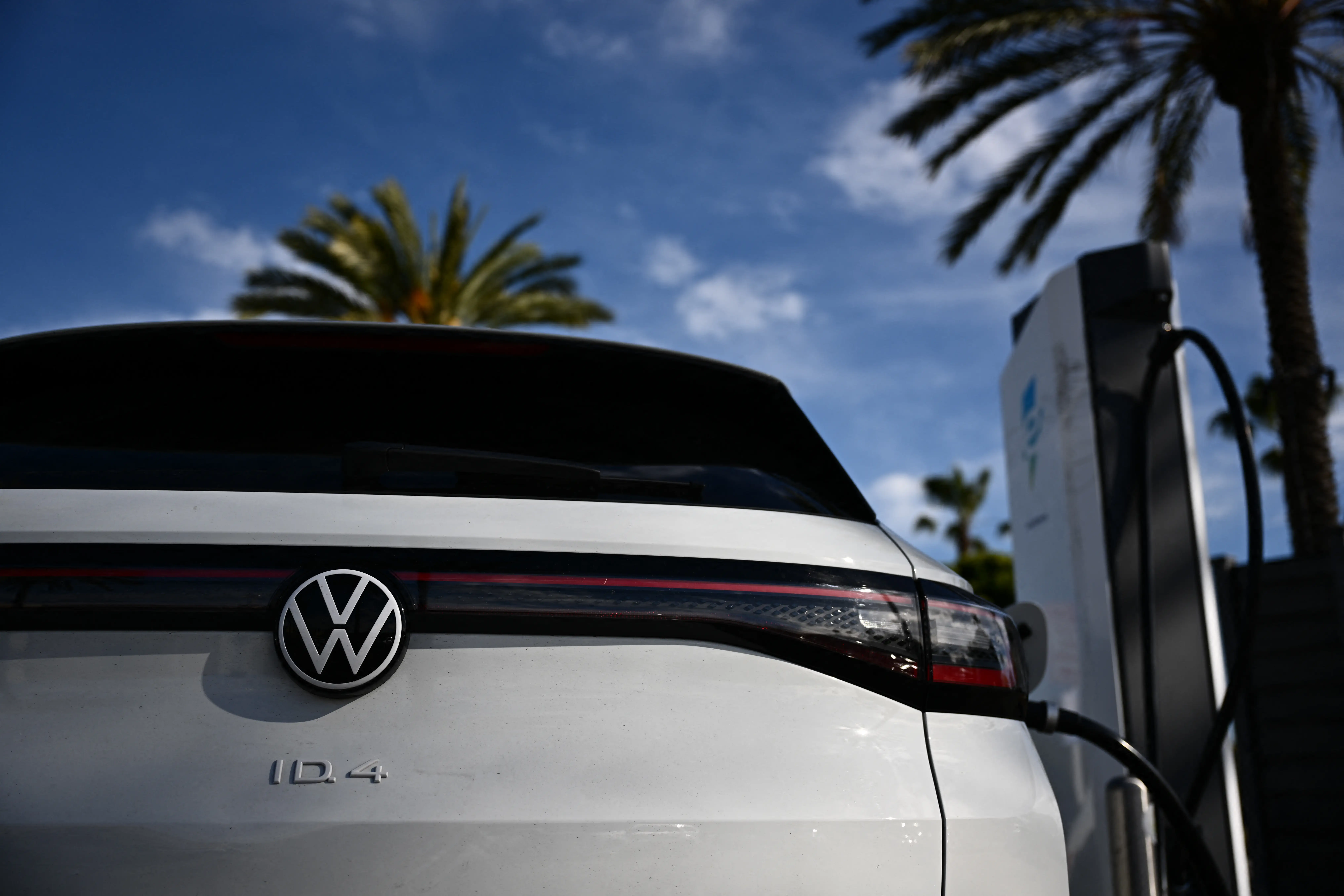 A white Volkswagen ID.4 is plugged into an Electrify America charge station next to two palm trees.