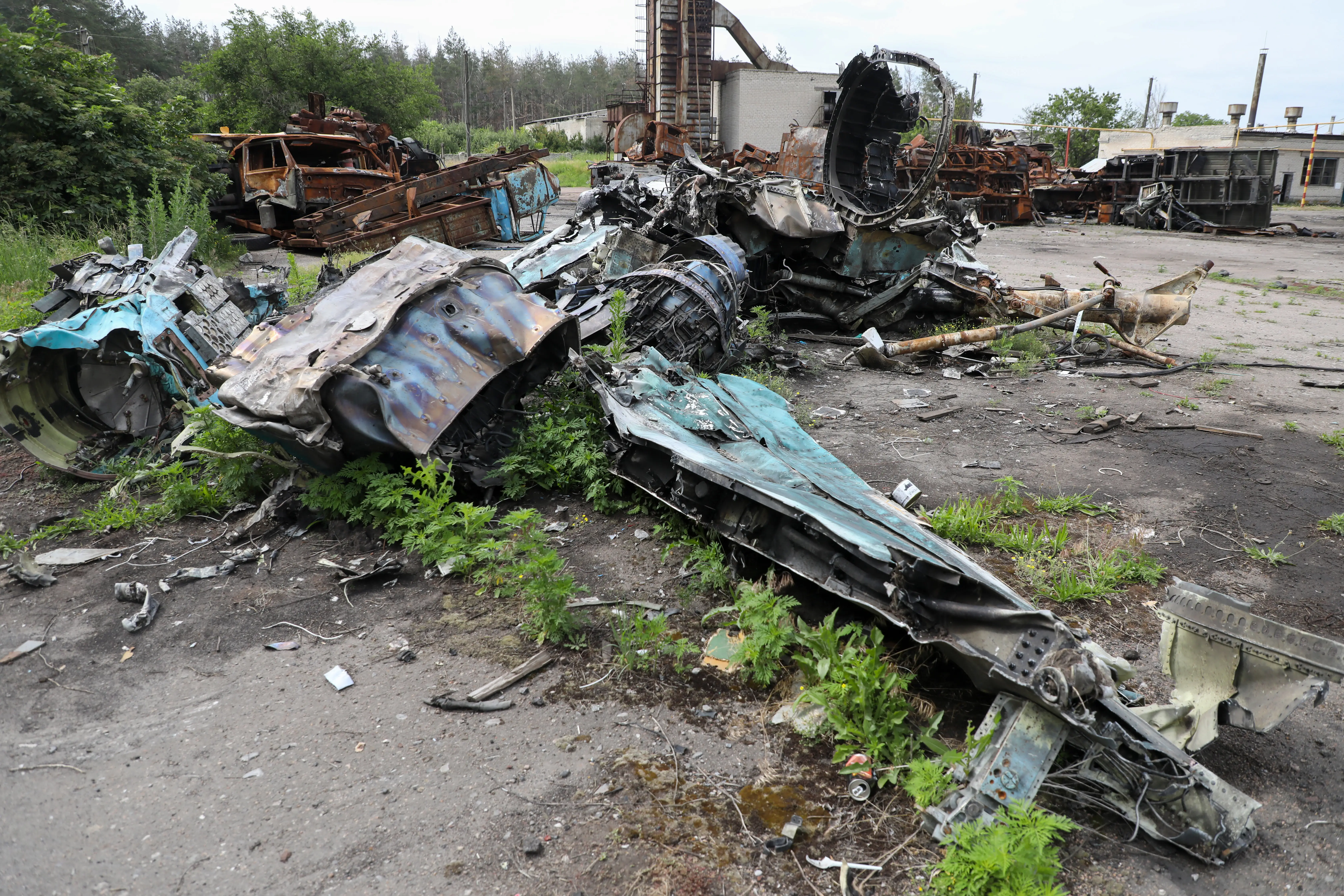 Destroyed remnants of a blue-colored jet on grassy tarmac with rusted vehicles behind