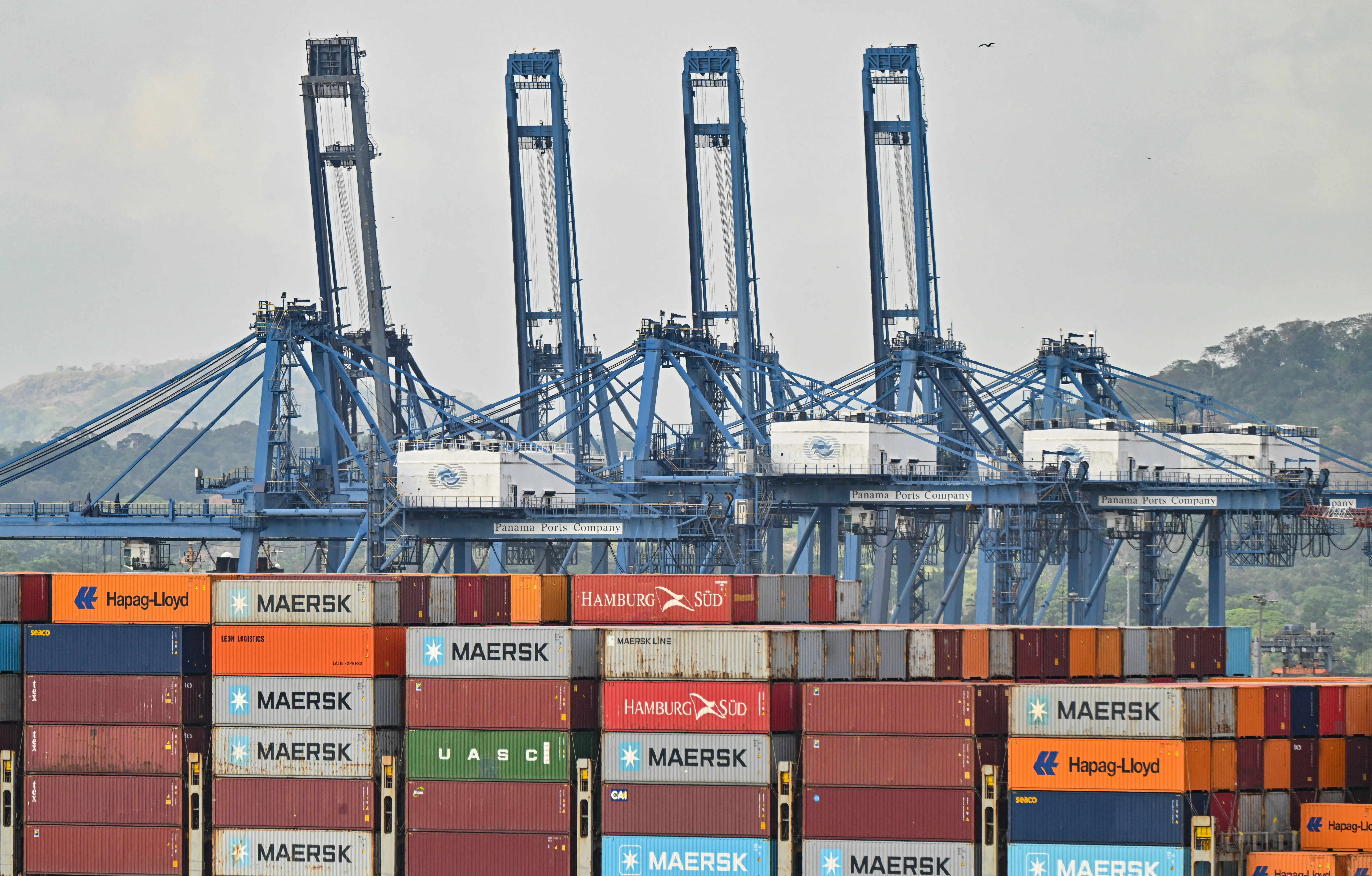 Shipping containers with names like Maersk, Hamburg Sud, and Hapag-Lloyd sit at a port as cranes stand in the background.