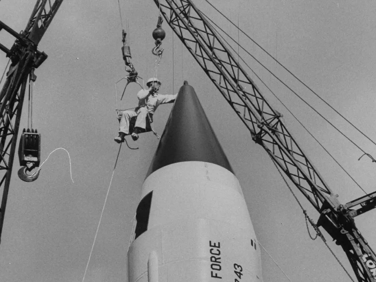 A construction worker attached to a crane touching the tip of a missile.