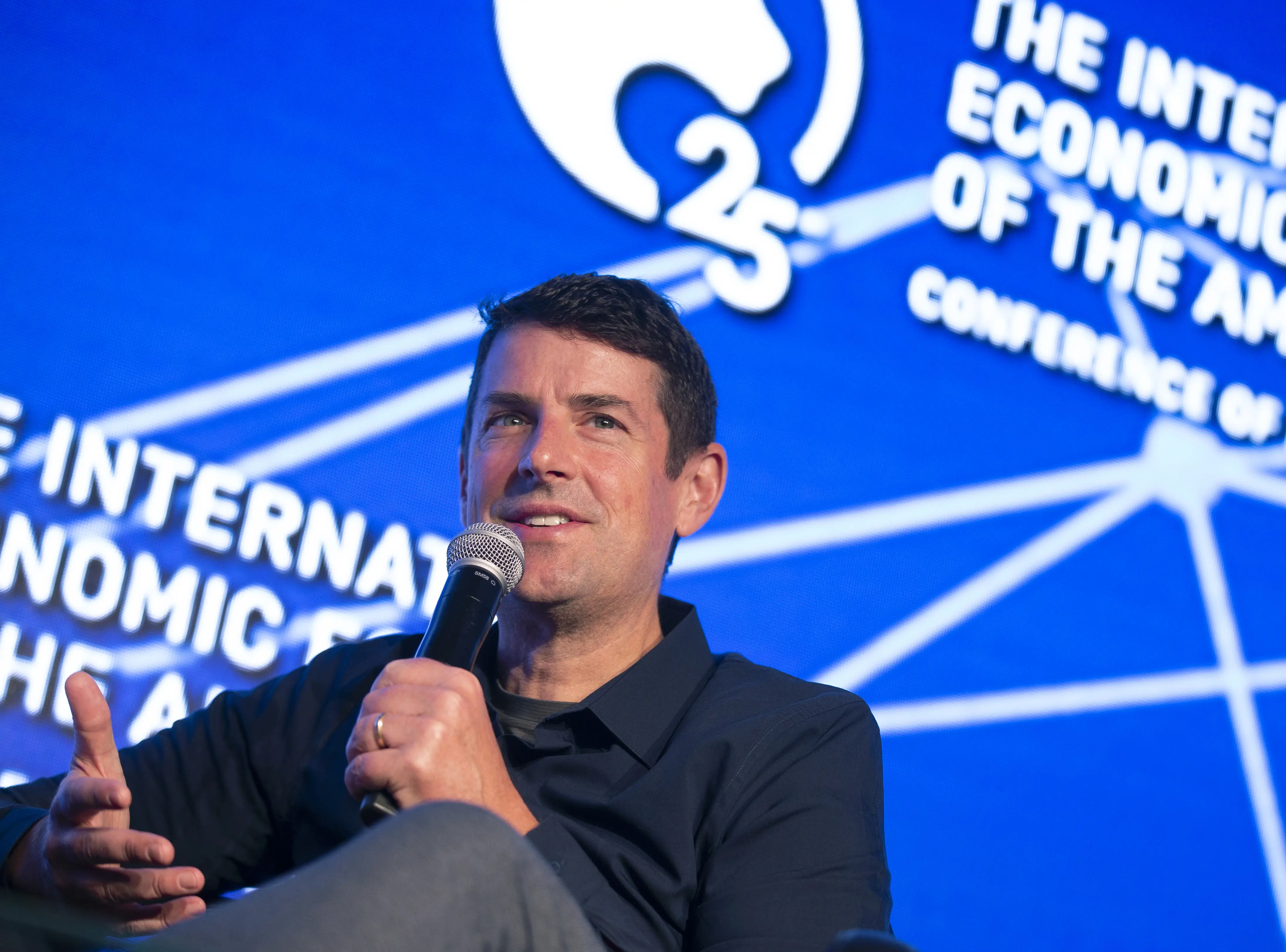 A man, Jon McNeill, holds a microphone onstage before a blue screen.