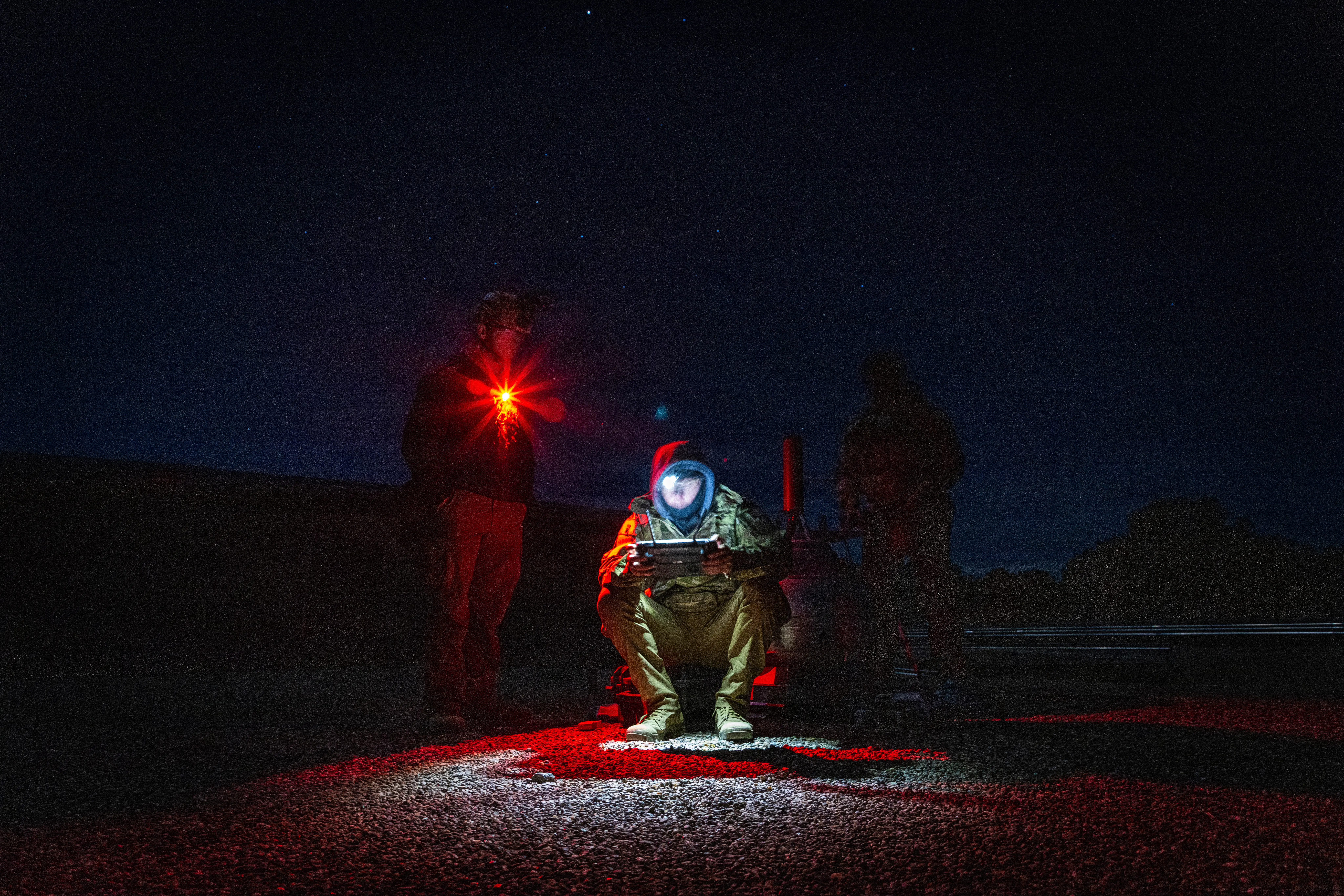 A soldier crouches looking at a tablet. Two soldiers stand behind him. It's nighttime.