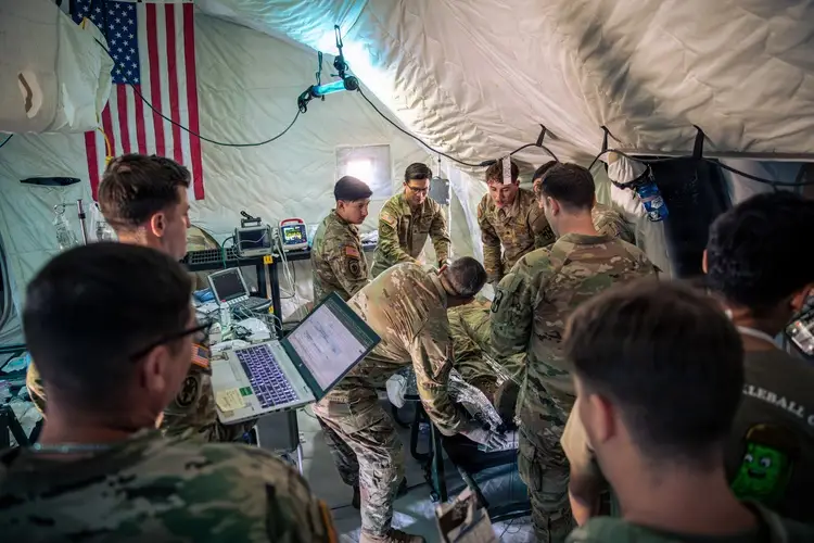 Soldiers stand around in a white tent. One soldier holds a laptop.