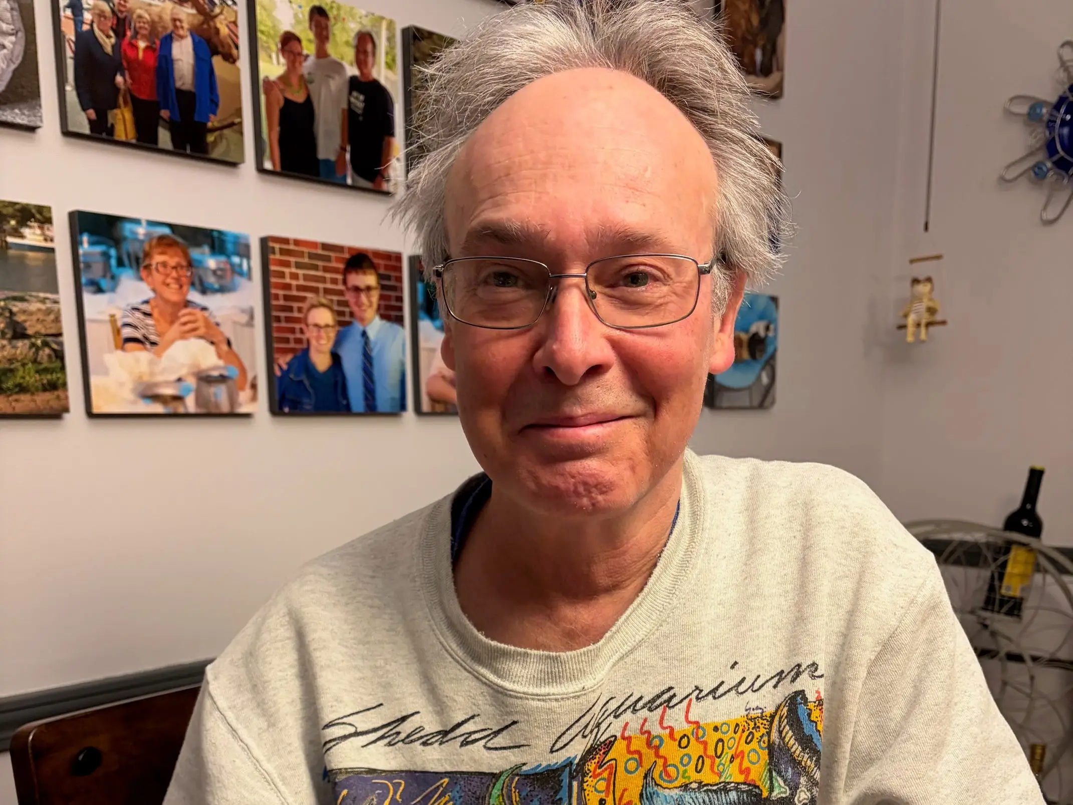 A man wearing glasses against a background of photos on the wall.