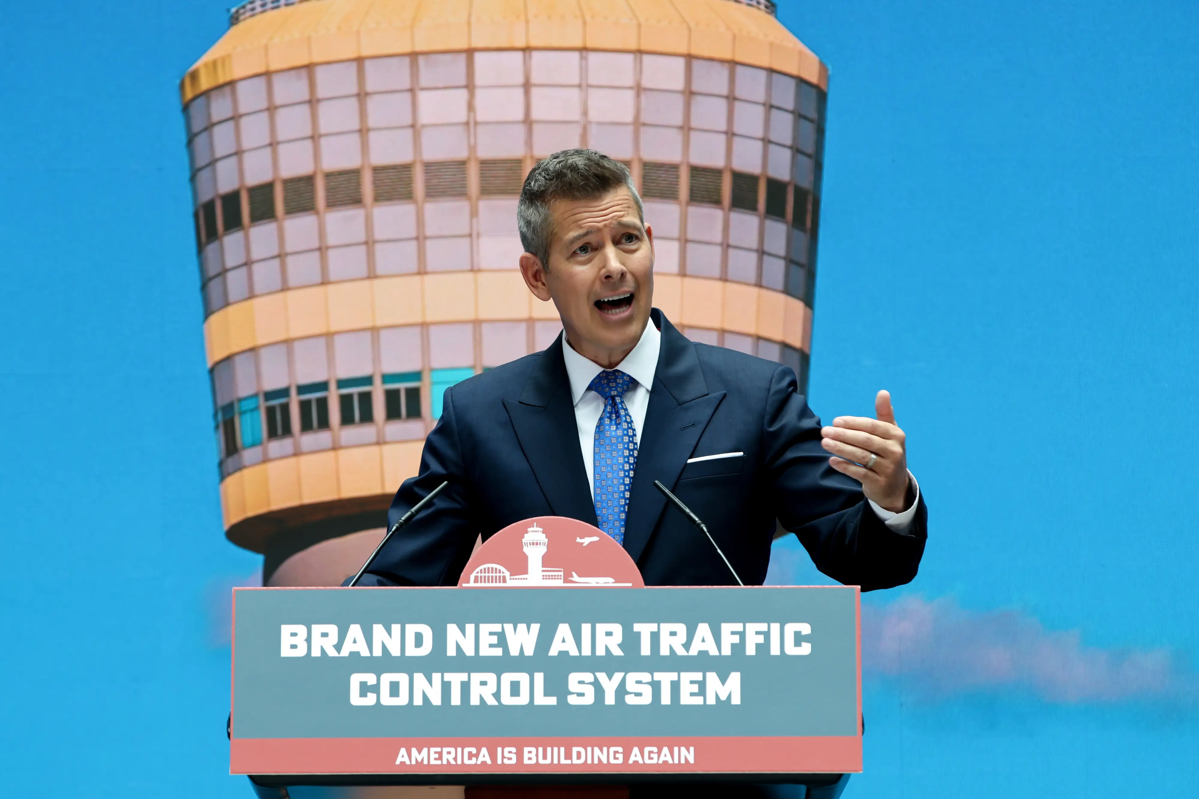 Transportation Secretary Sean Duffy speaks at a press conference behind a podium with an air traffic control tower pictured behind him.