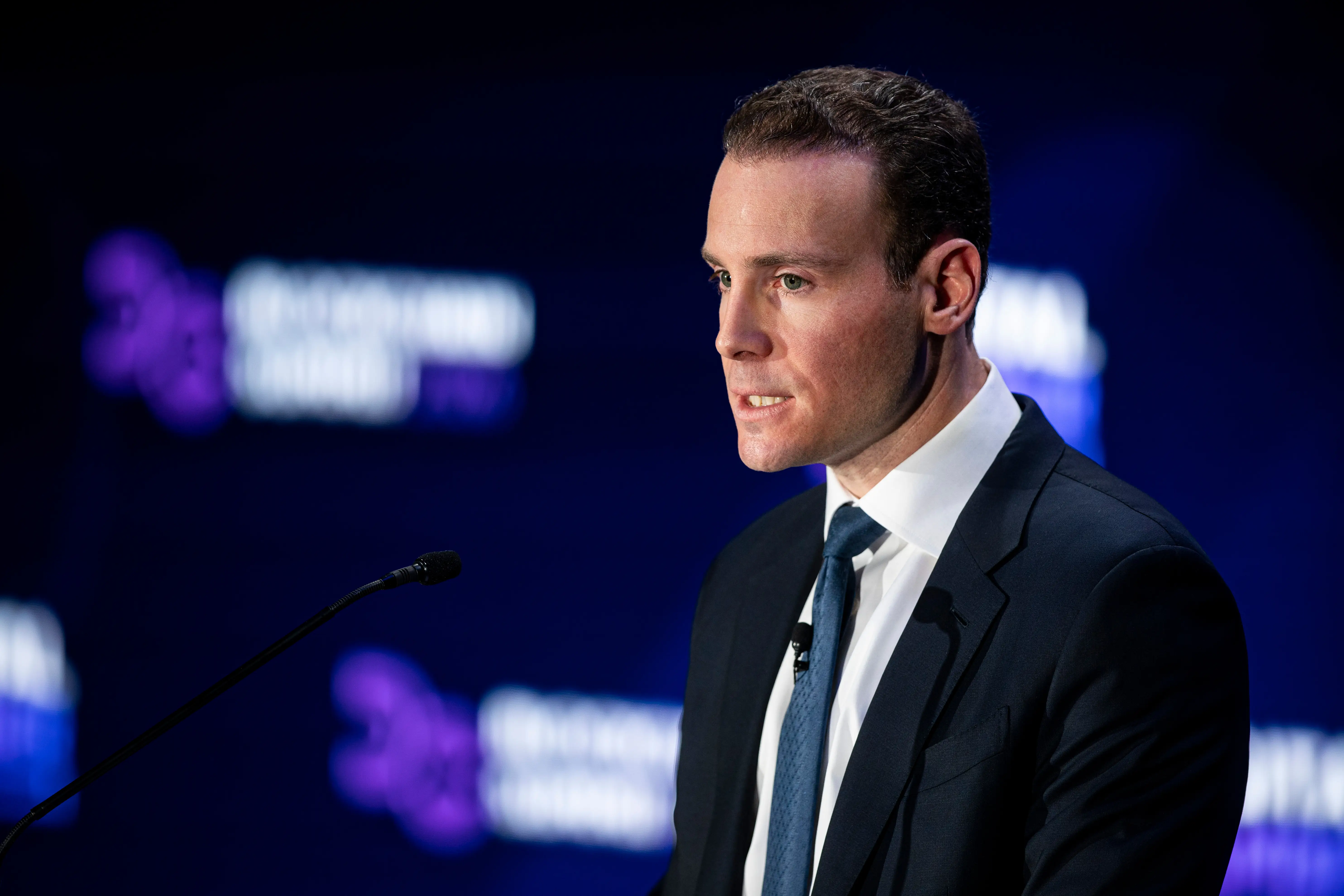 Michael Selig, in a black suit and blue tie, speaks from behind a podium against a purple background