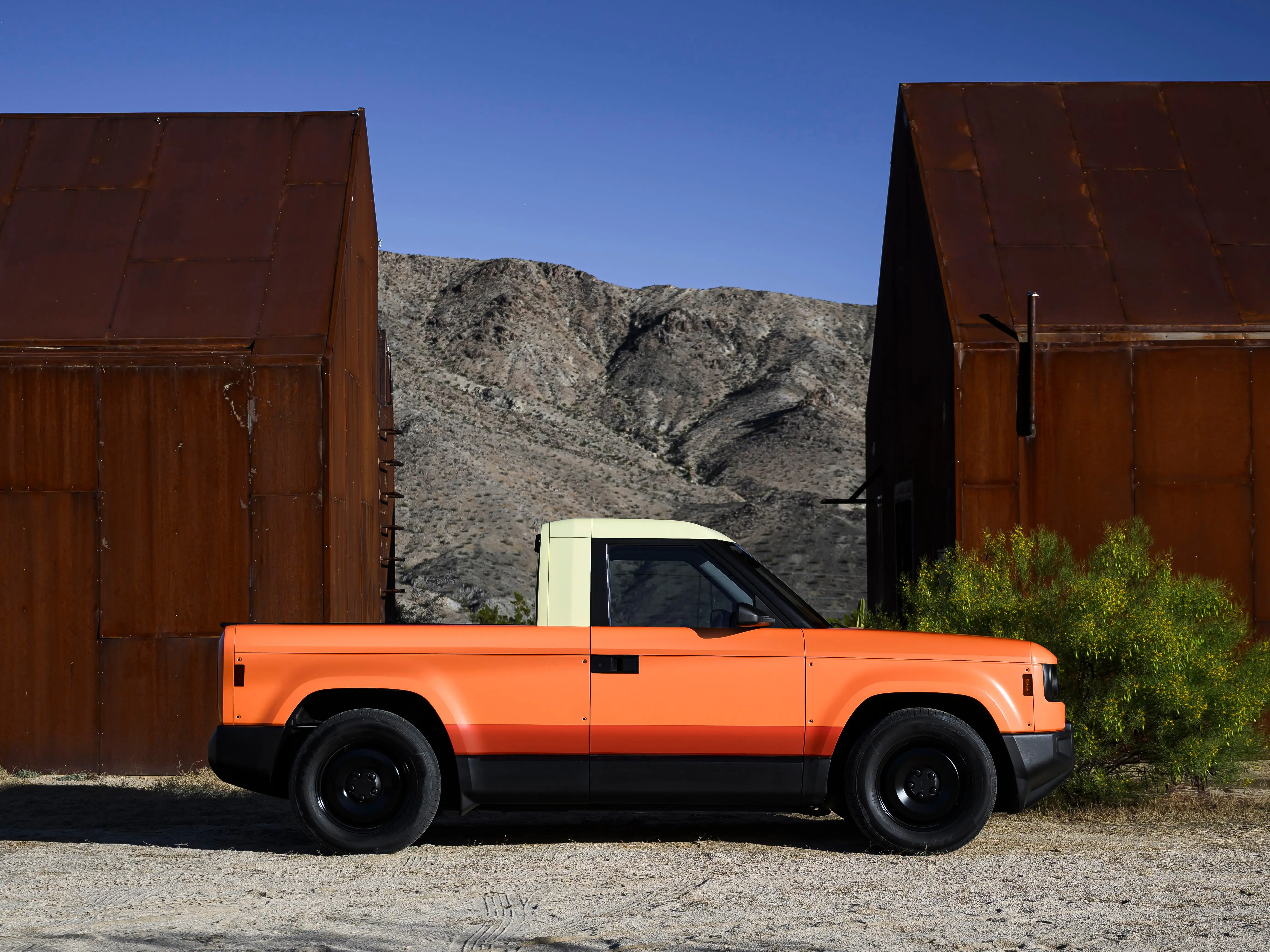 A Slate Truck, wrapped in orange with a white top, is parked next to two rust-brown buildings on a clear day.