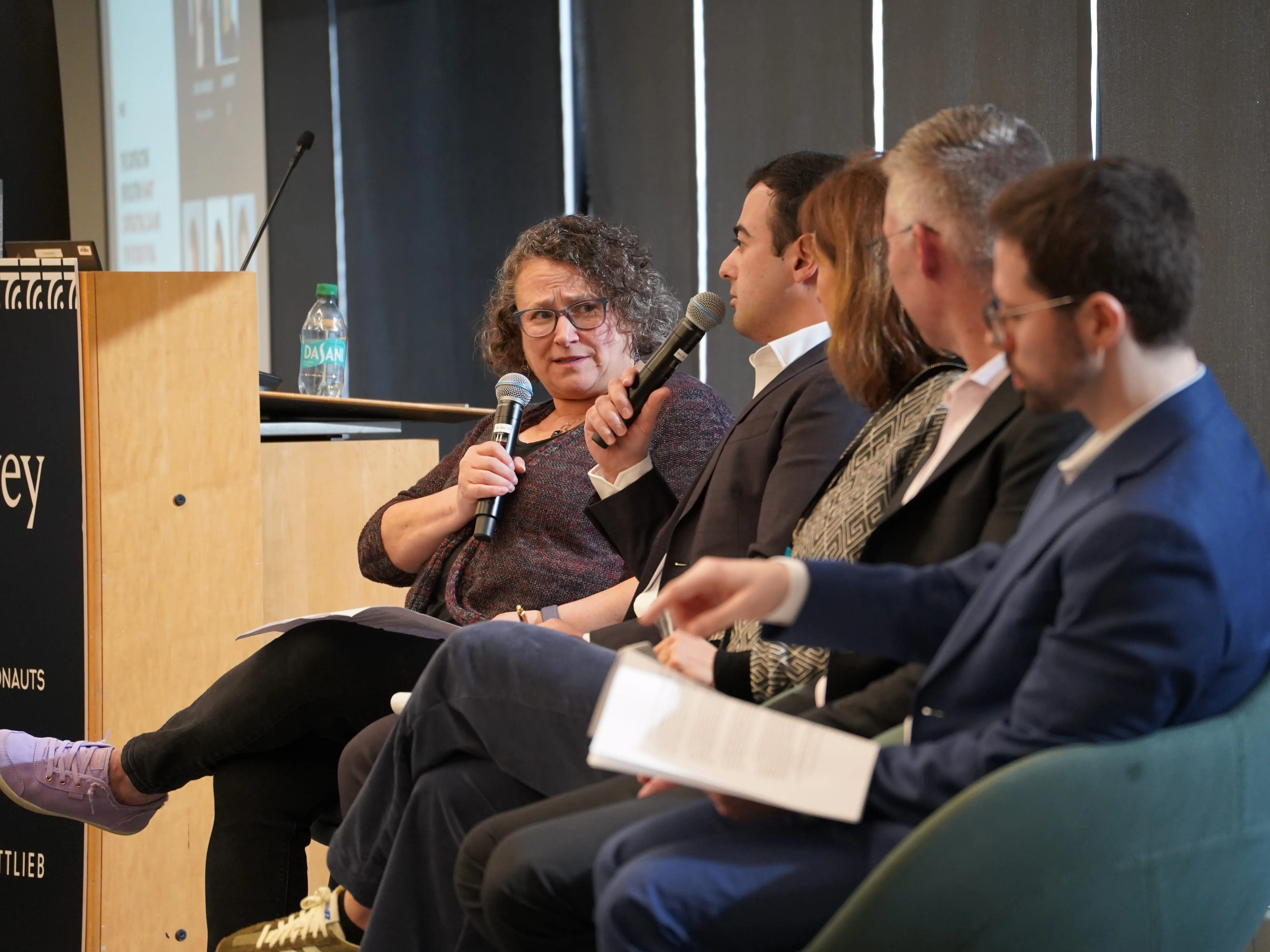 A woman holding a microphone speaks to a seated panel of businesspeople during a conference discussion.