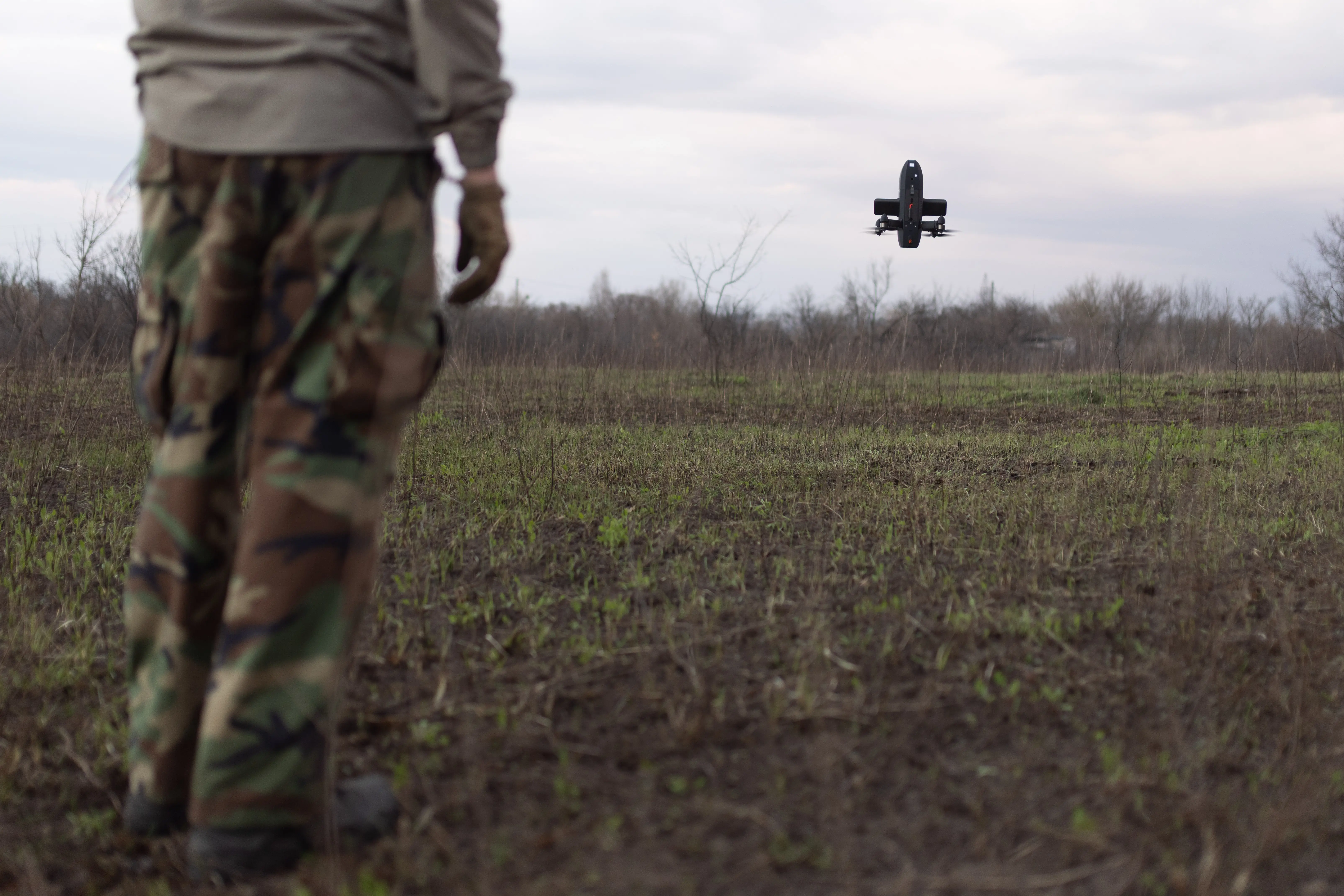 The legs of a figure wearing camouflage in the foreground on a grassy and muddy field with a small interceptor drone in the air