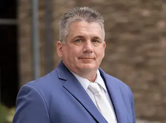 Bill Lewis poses for the camera wearing a blue suit, white dress shirt, and white tie.