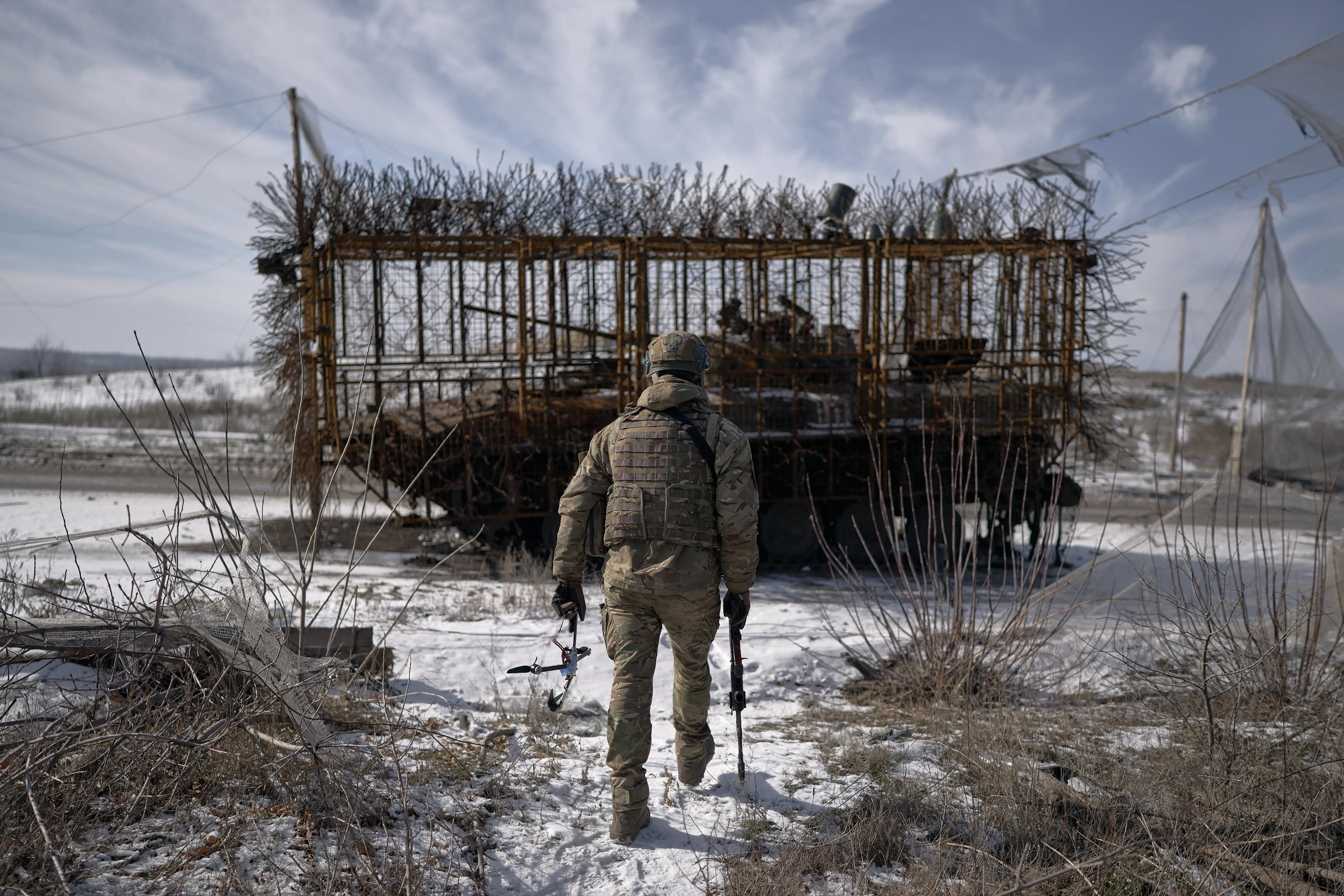 A figure in camouflage walks with his back turned on a snowy ground towards an armored vehicle covered in a spiky cage