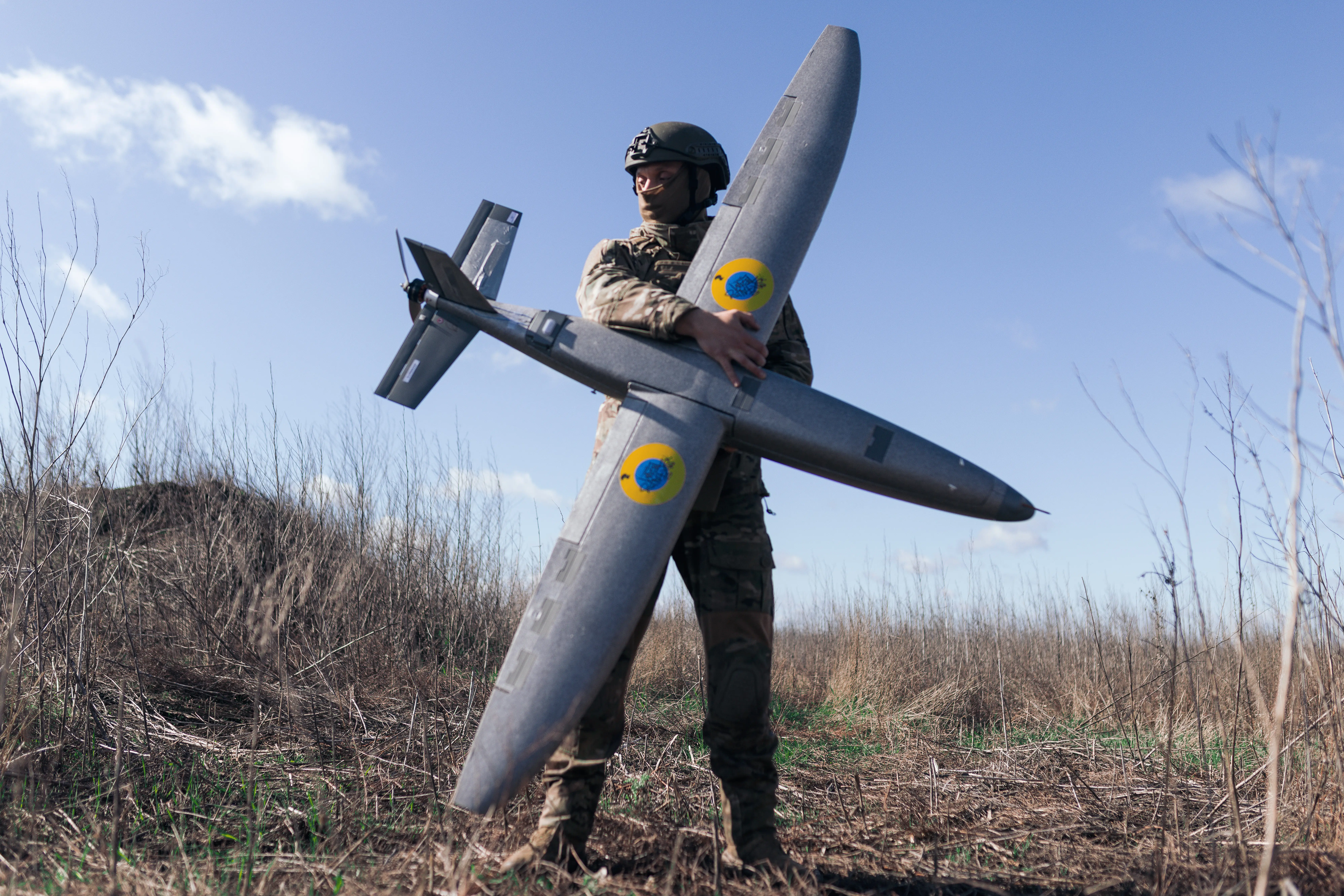 A man in camouflage and a helmet stands holding a large grey drone in a sunny field