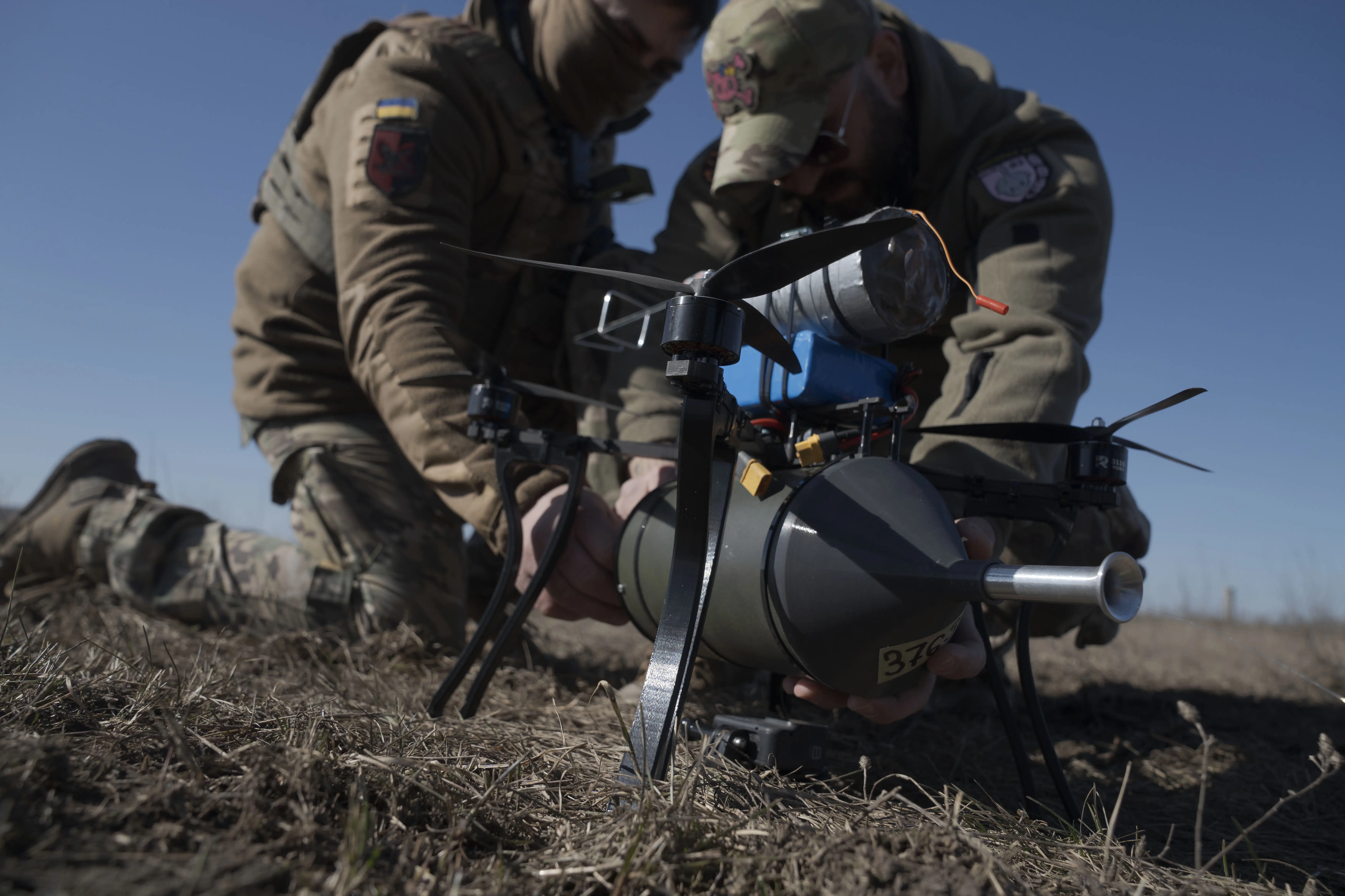 Two men in khaki kneel in a sunny field working on a dark-colored drone