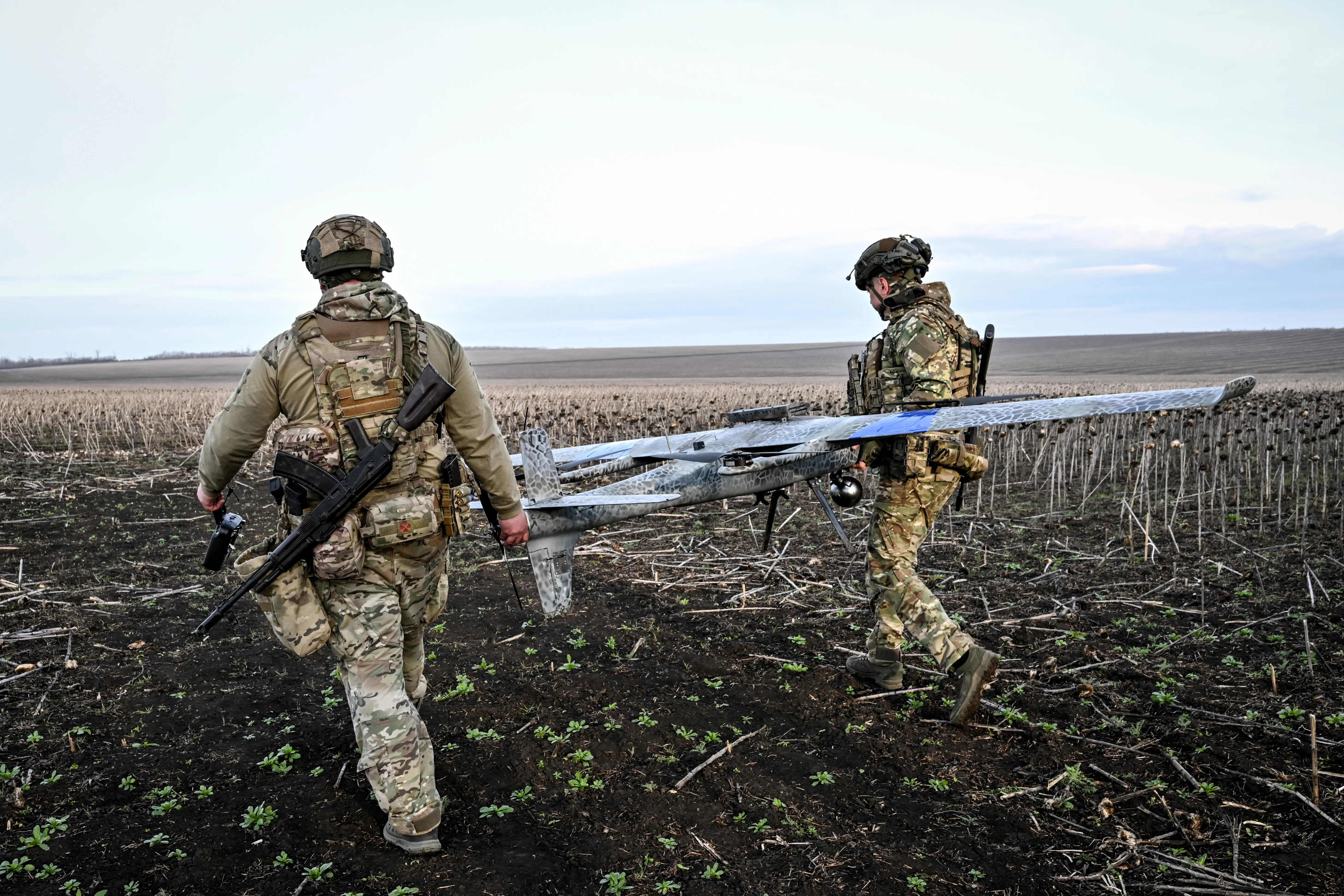 Two figures in camouflage gear walk on a brown field holding a flat grey large drone