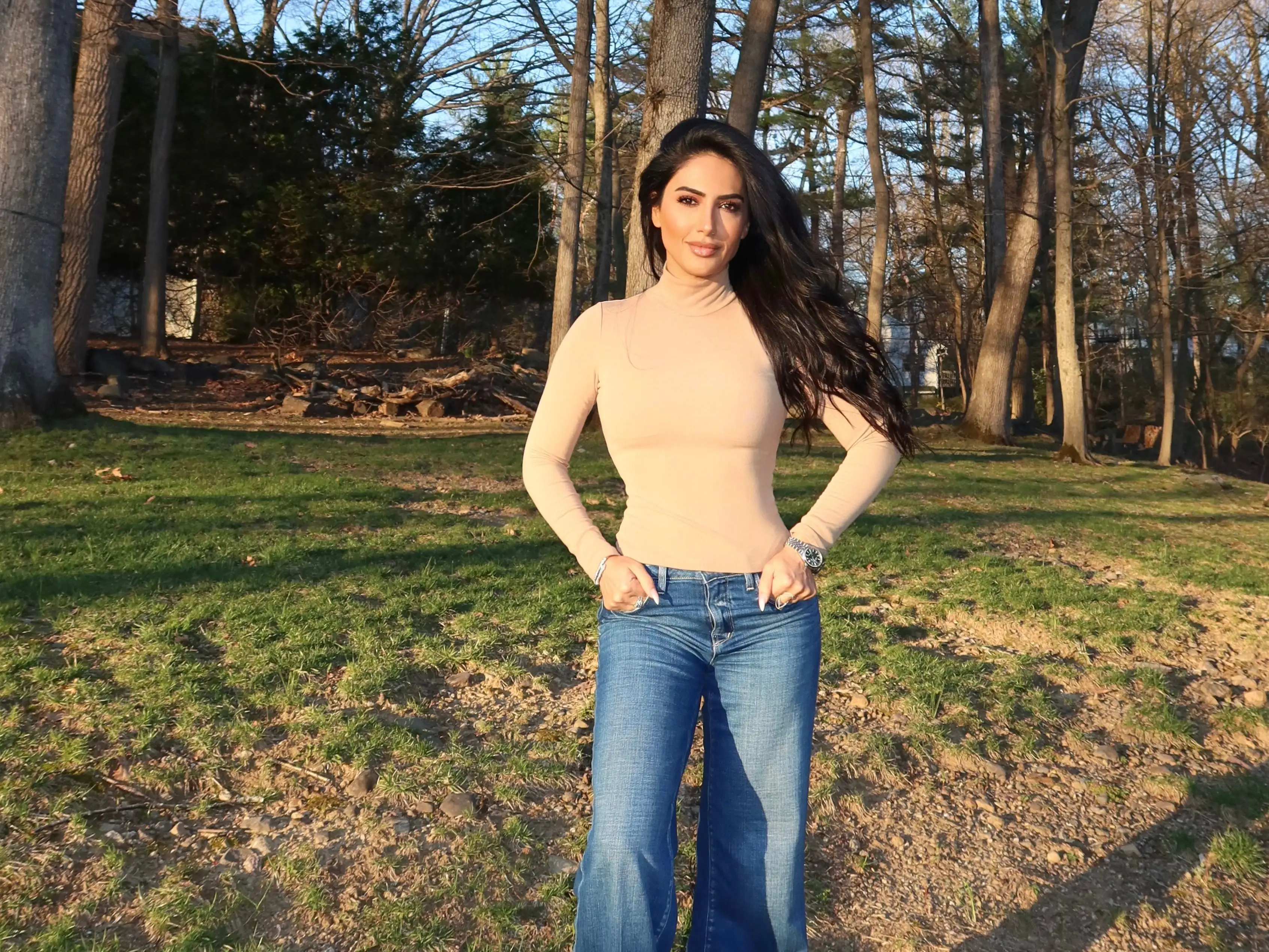 A woman wearing jeans and a beige polo neck sweater standing in front of trees.