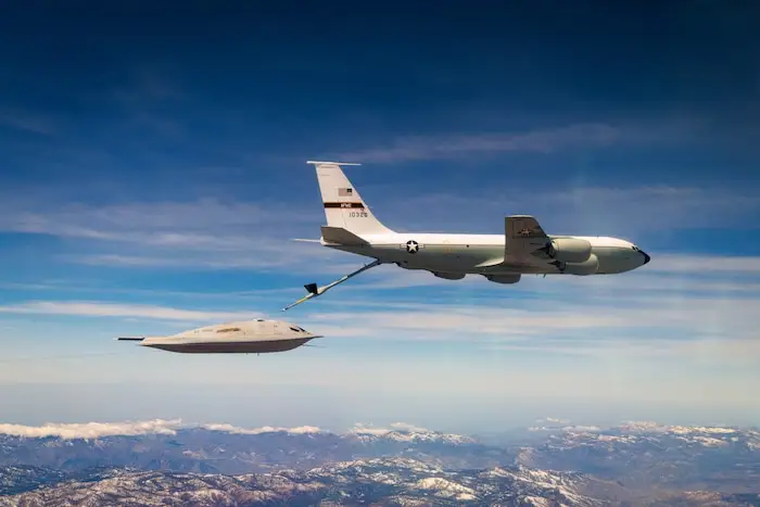 A B-21 Raider flies behind an aerial refueling aircraft in a mountainous landscape.