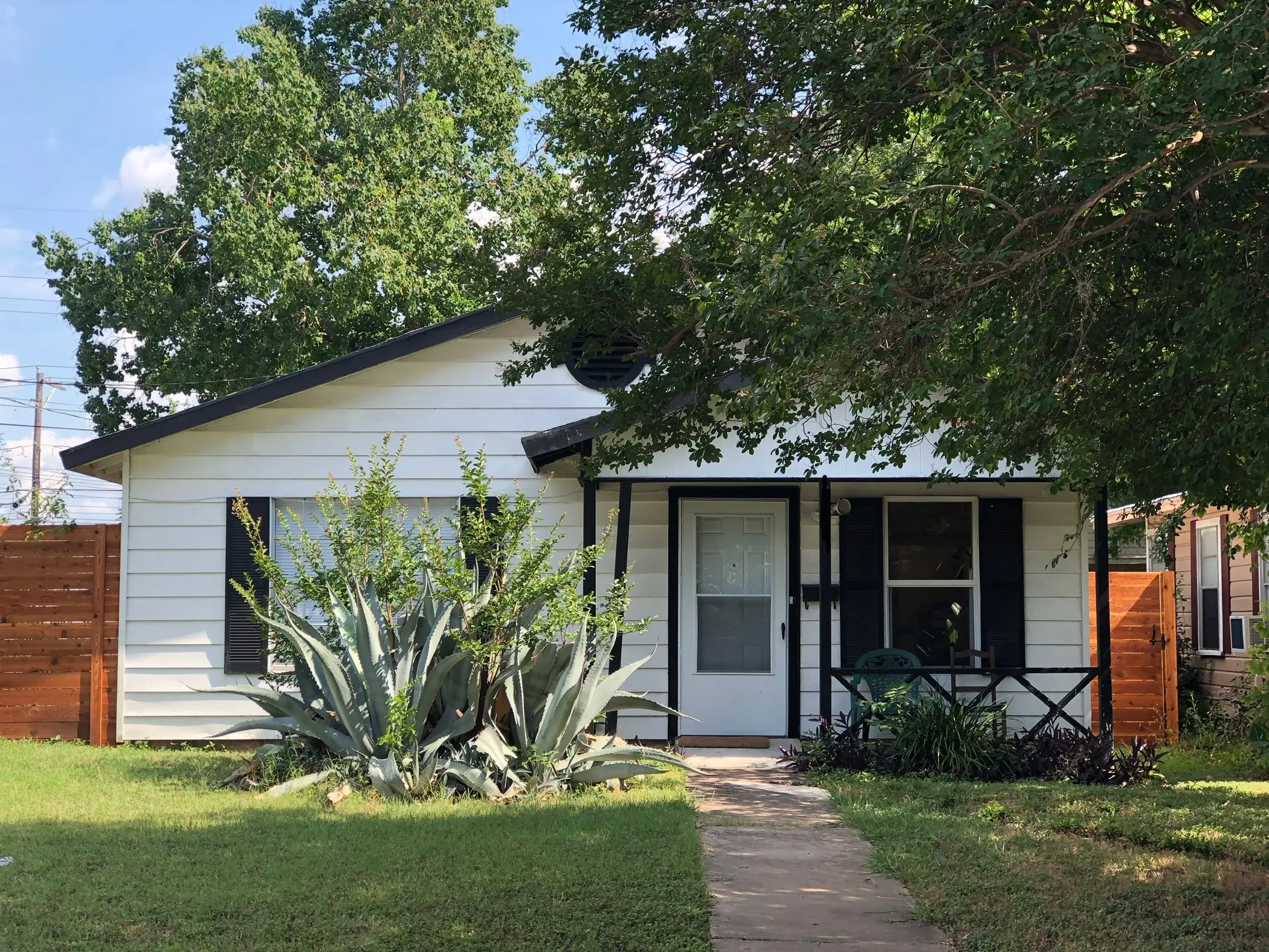 A modest-sized white home sits underneath two tall trees.