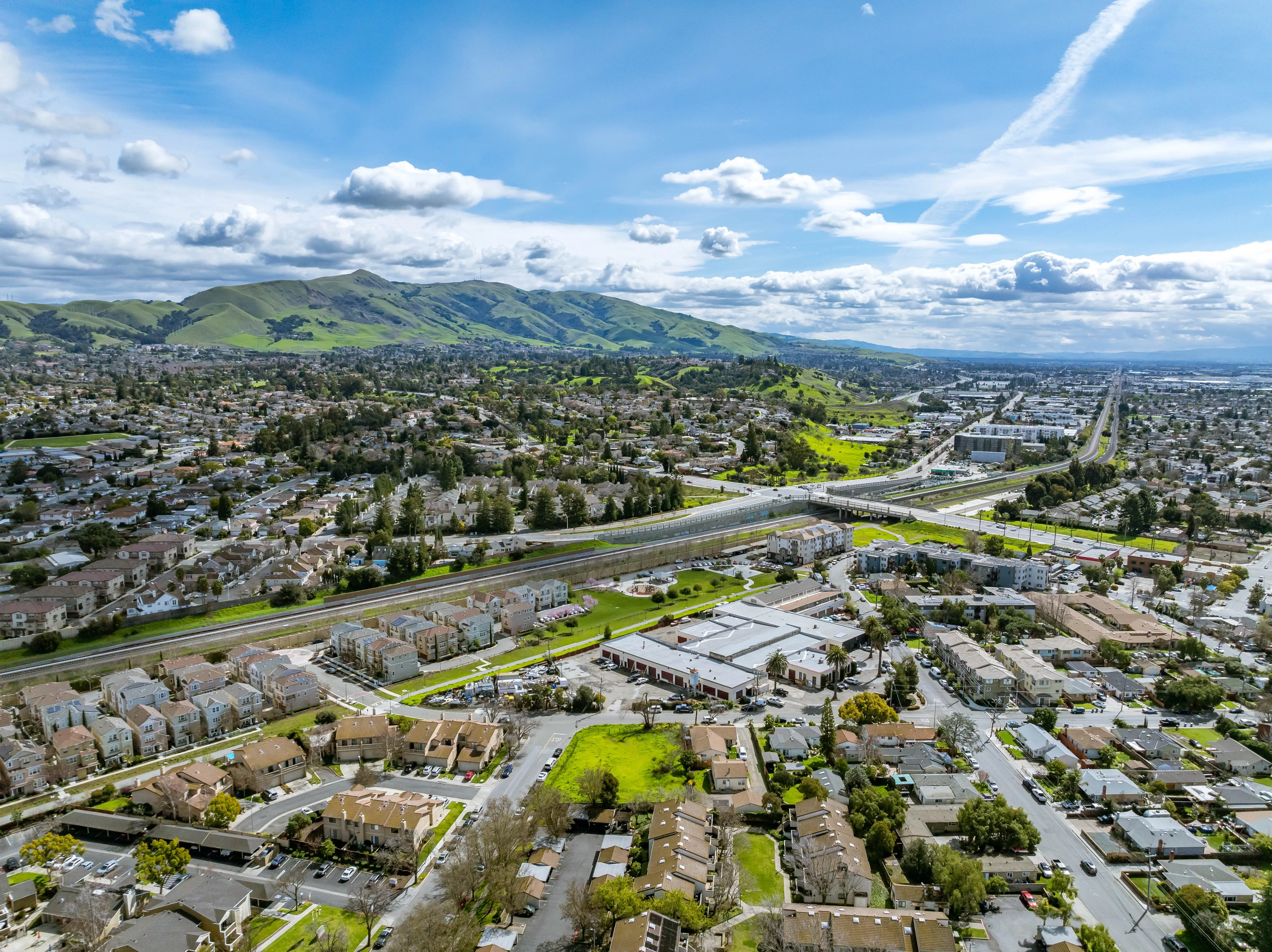 Drone images showcasing residential neighborhoods in Fremont, California, with lush green hills in the background and a mix of townhomes, single-family homes, and commercial buildings. Ideal for theme