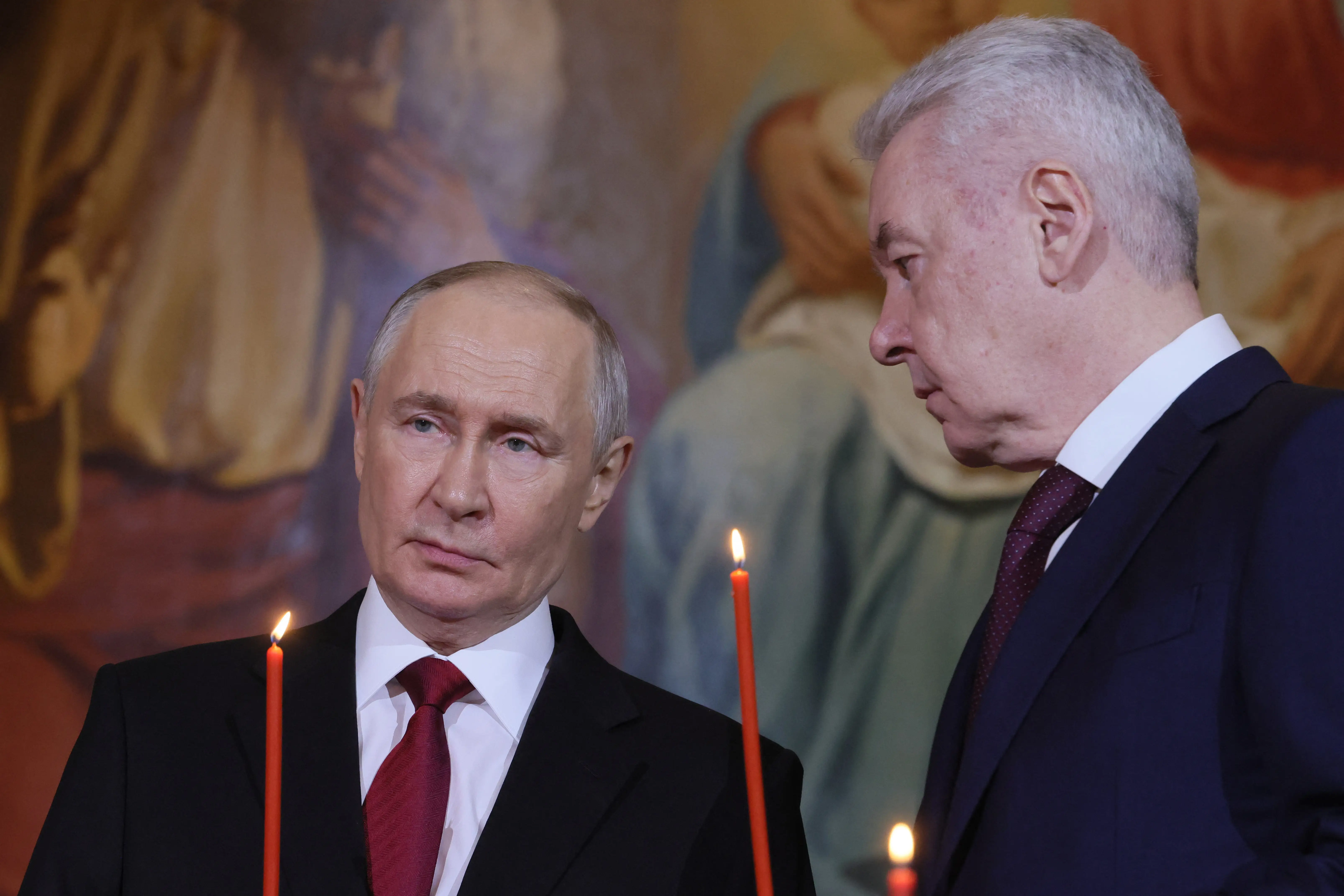 Sergey Sobyanin looks at Vladimir Putin as both hold candles.