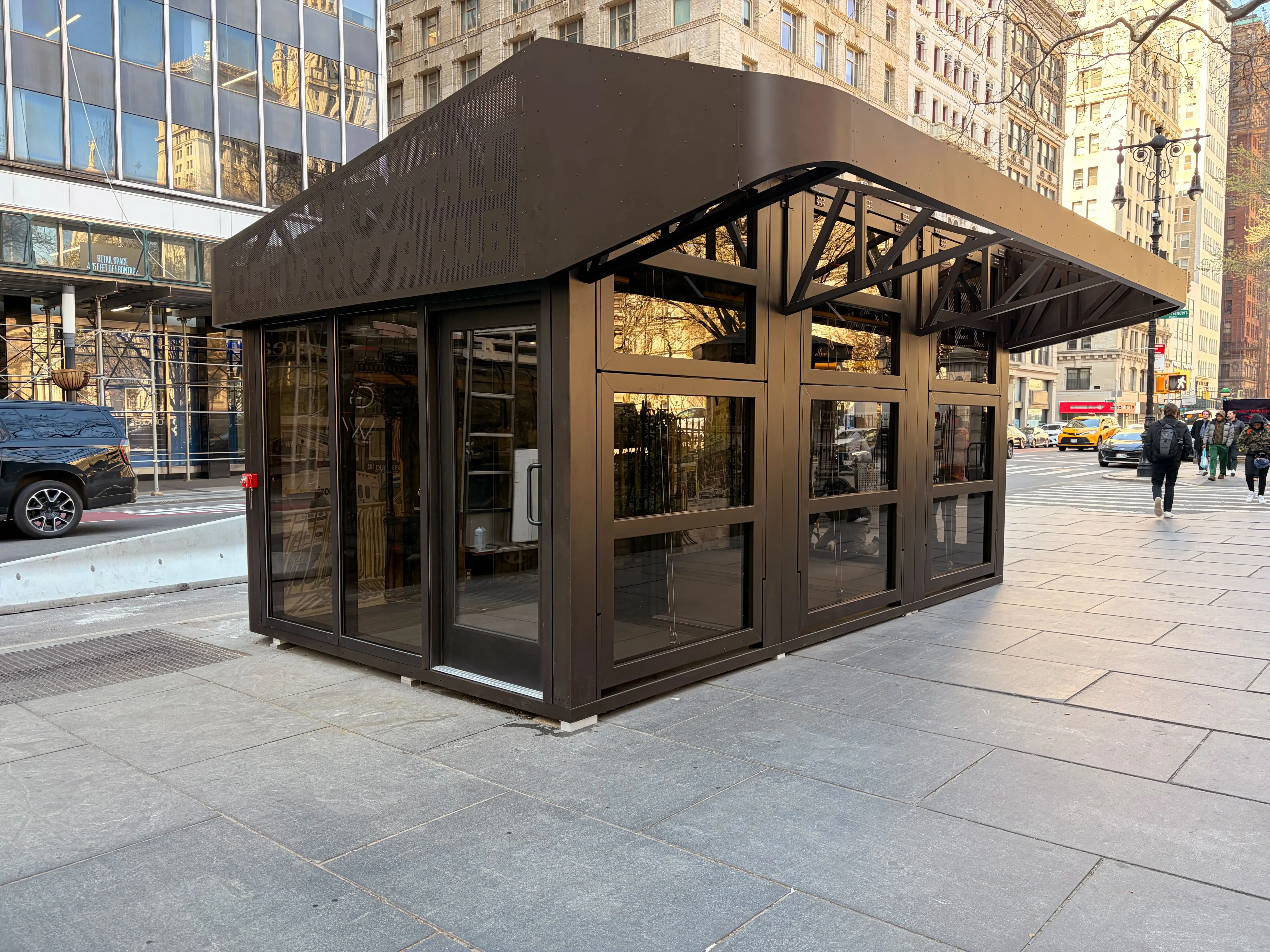 A dark brown metal building rests on a New York City sidewalk outside the City Hall.