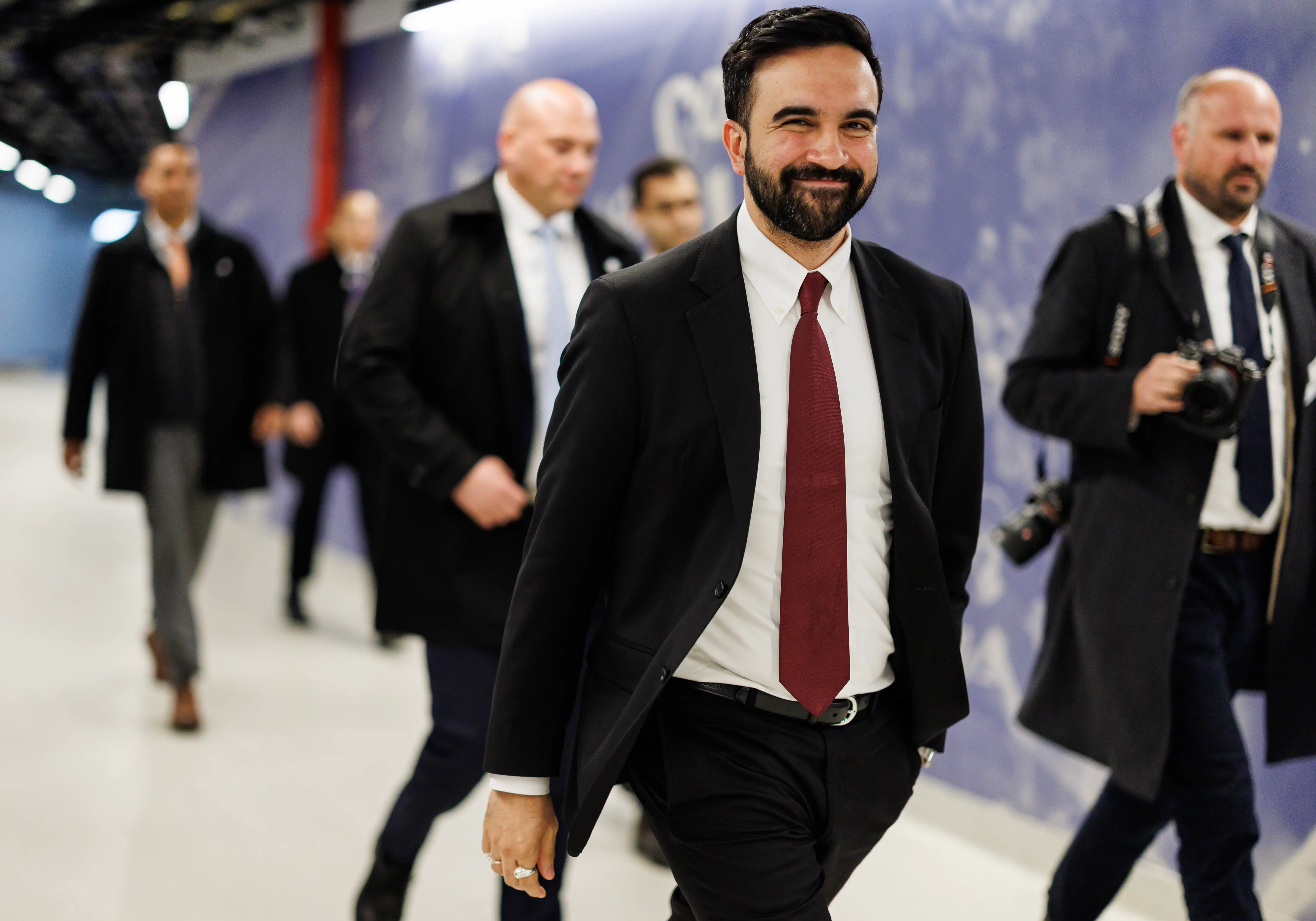 Zohran Mamdani grins toward the camera, flanked by photographers.