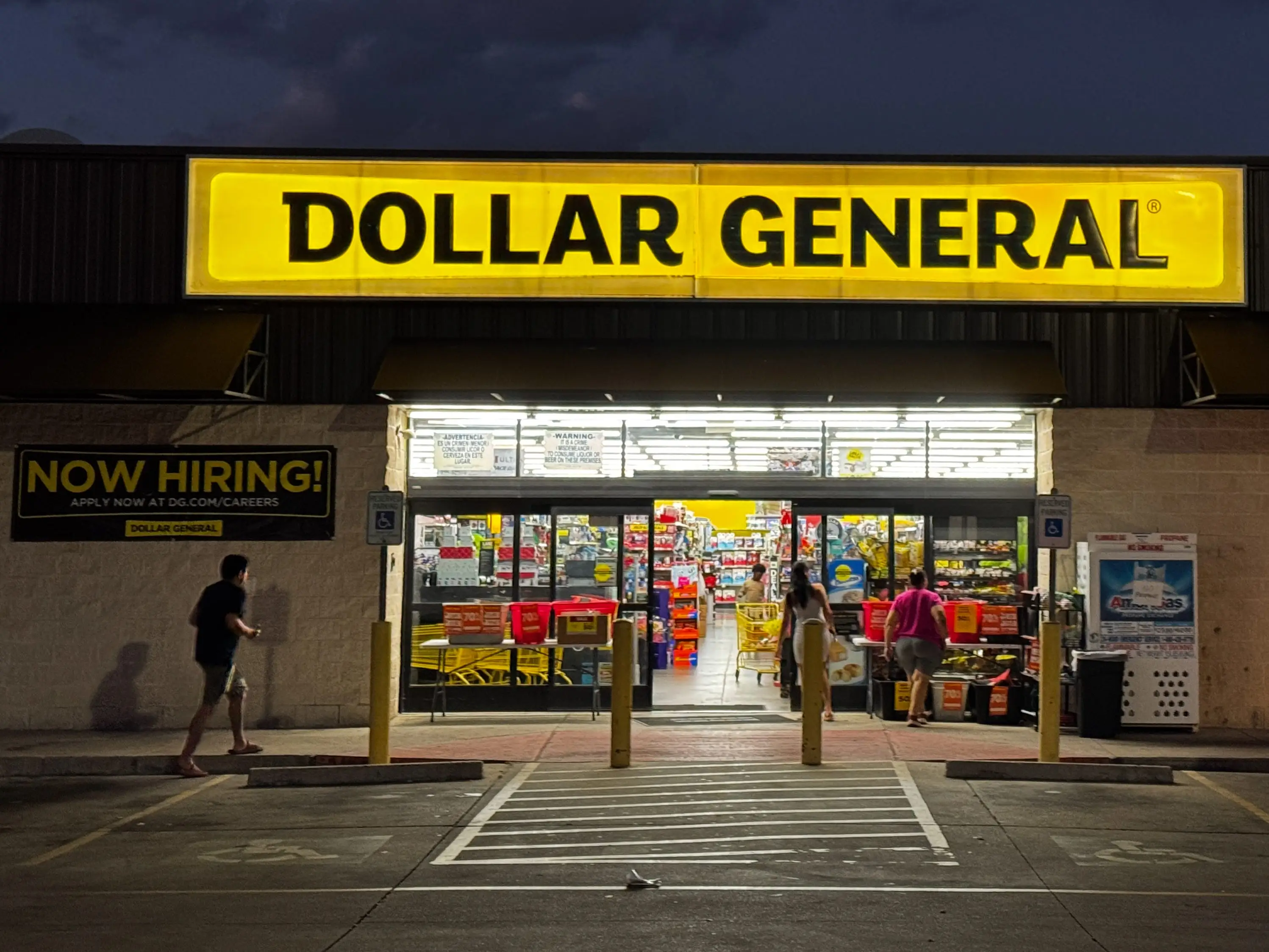 A Dollar General store is seen at night as customers walk in the main entrance from the parking lot.