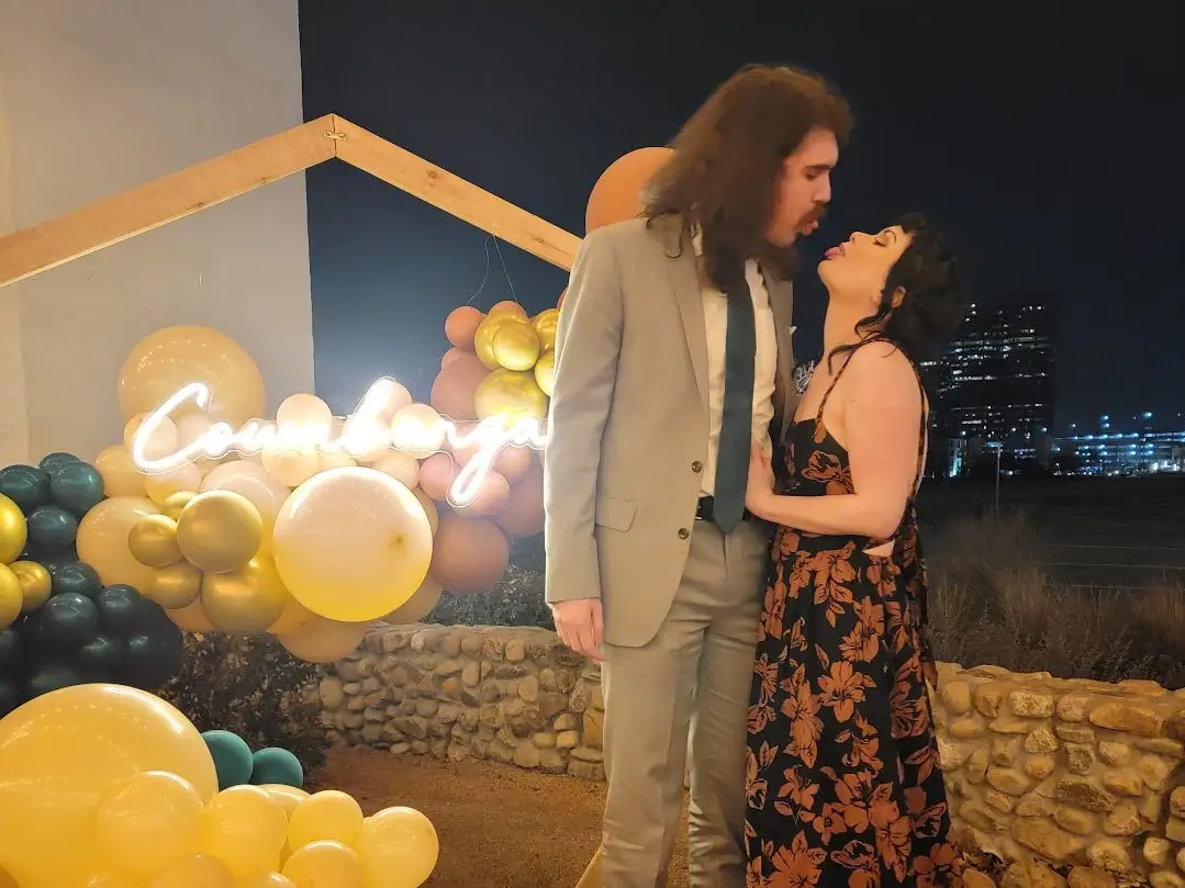 A man and woman stick their tongue out at each other, standing in front of a sign.