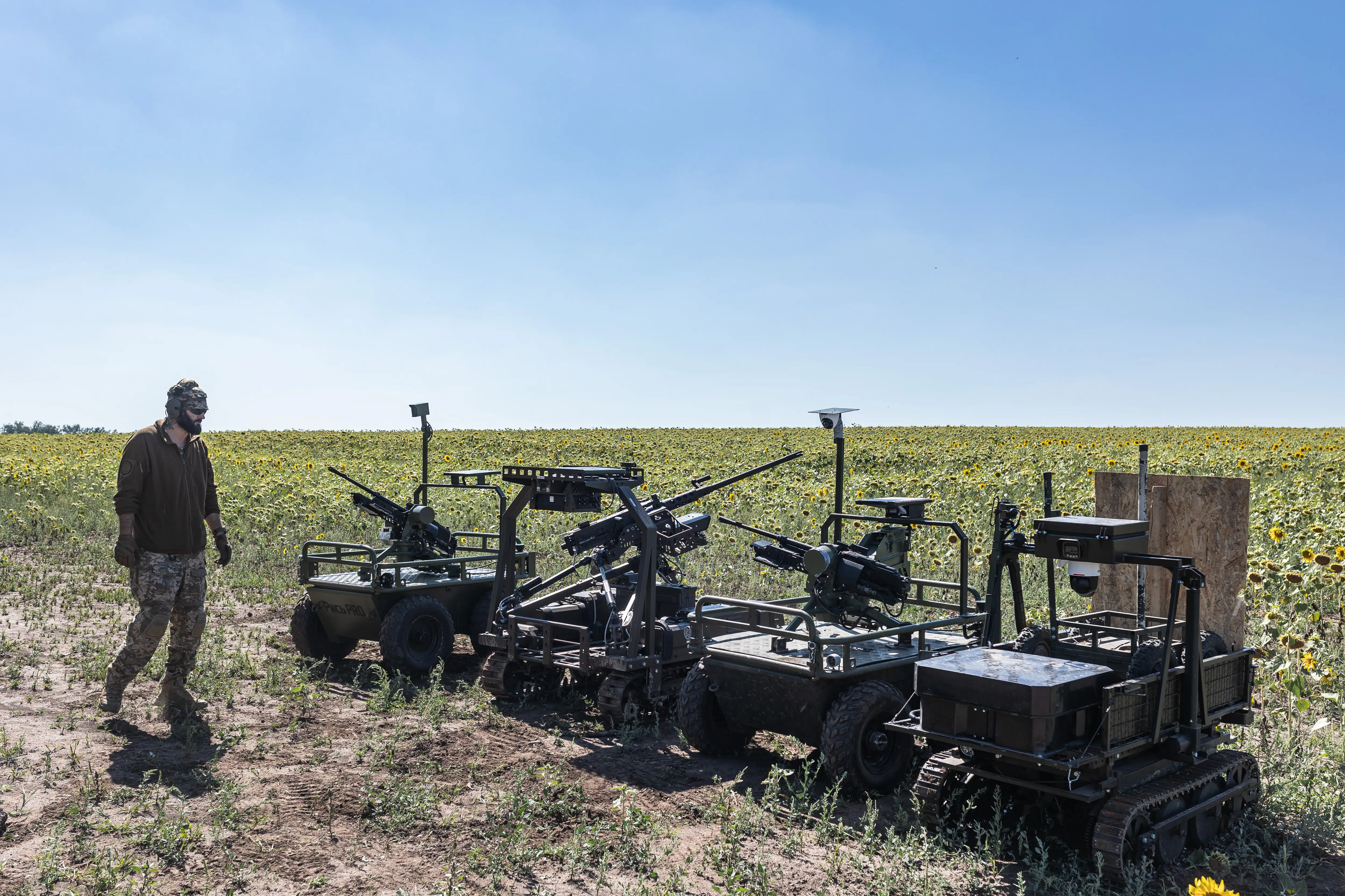 A man in khaki stands beside multiple green-colored robots in a sunflower field under a blue sky