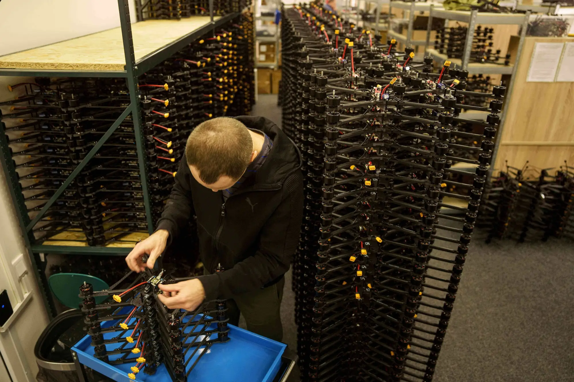 A man bends over a small stack of drones on a blue surface with stacks of small black drones behind him in a room