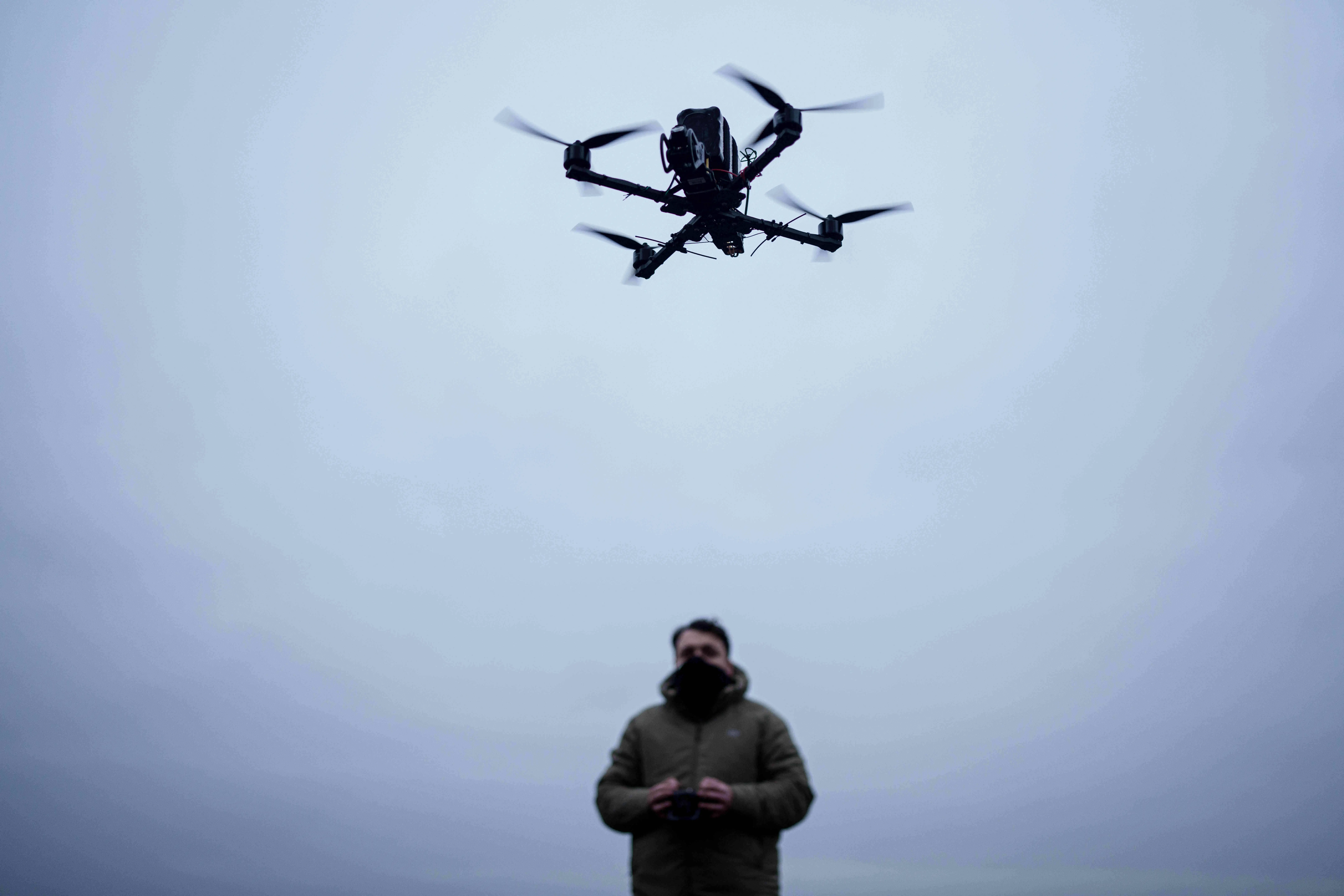 A man wearing a face covering and in a khaki coat stands in a cloudy sky holding a controller with a black quadcopter drone flying above him