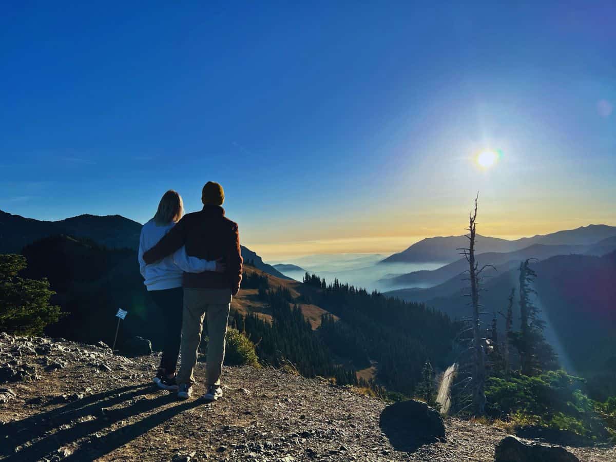 Scott and his wife drape their arms around each other while looking out at the sunrise over the mountains in Olympic National Park.