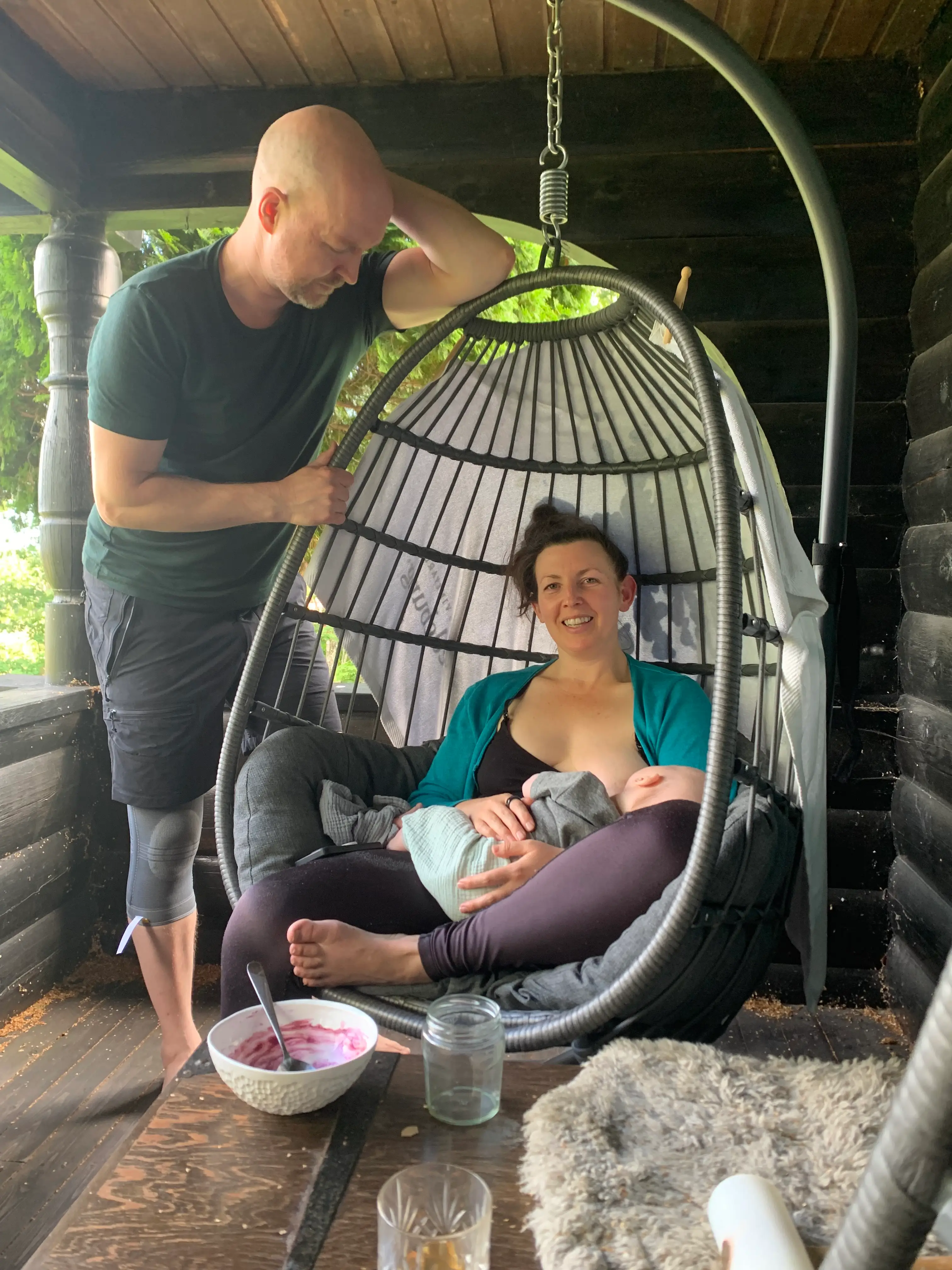 Woman smiling with child in lap in egg chair with man standing next to her, leaning on chair