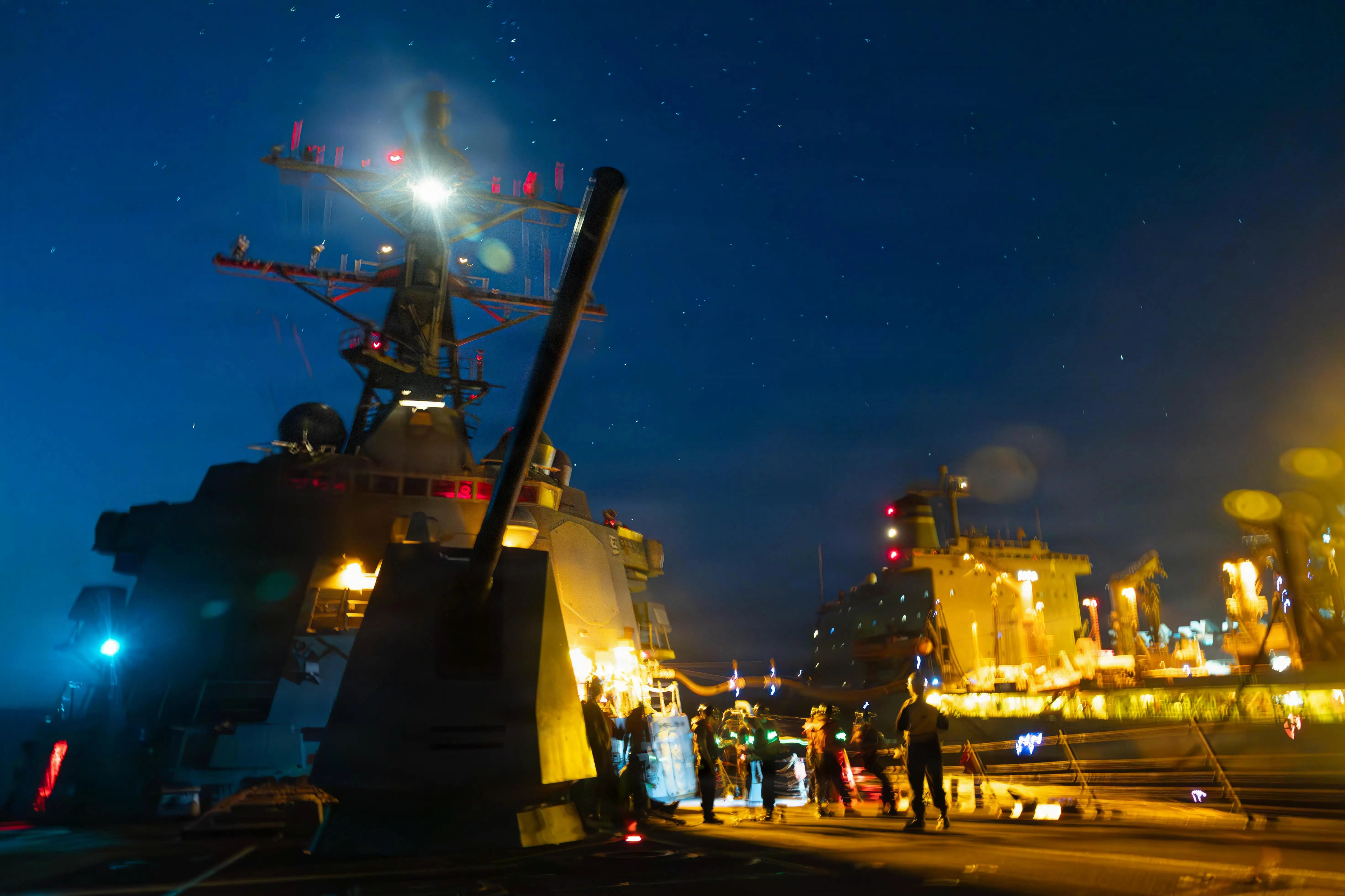 The front of the USS Spruance is seen at night with another vessel next to it conducting an at-sea refueling.