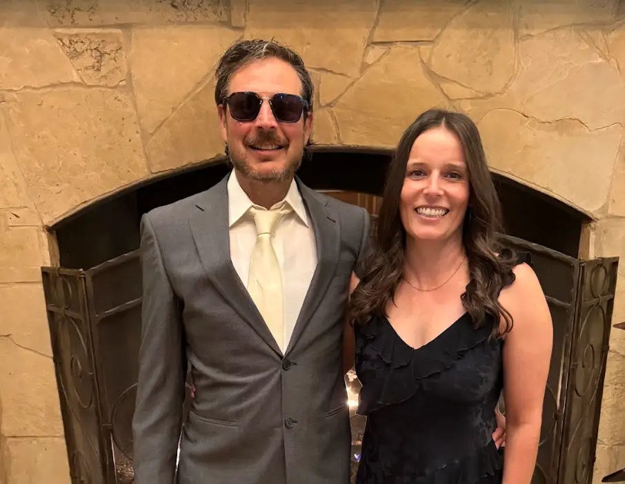 The writer and his wife dressed up for an event and standing in front of a fireplace in a house.
