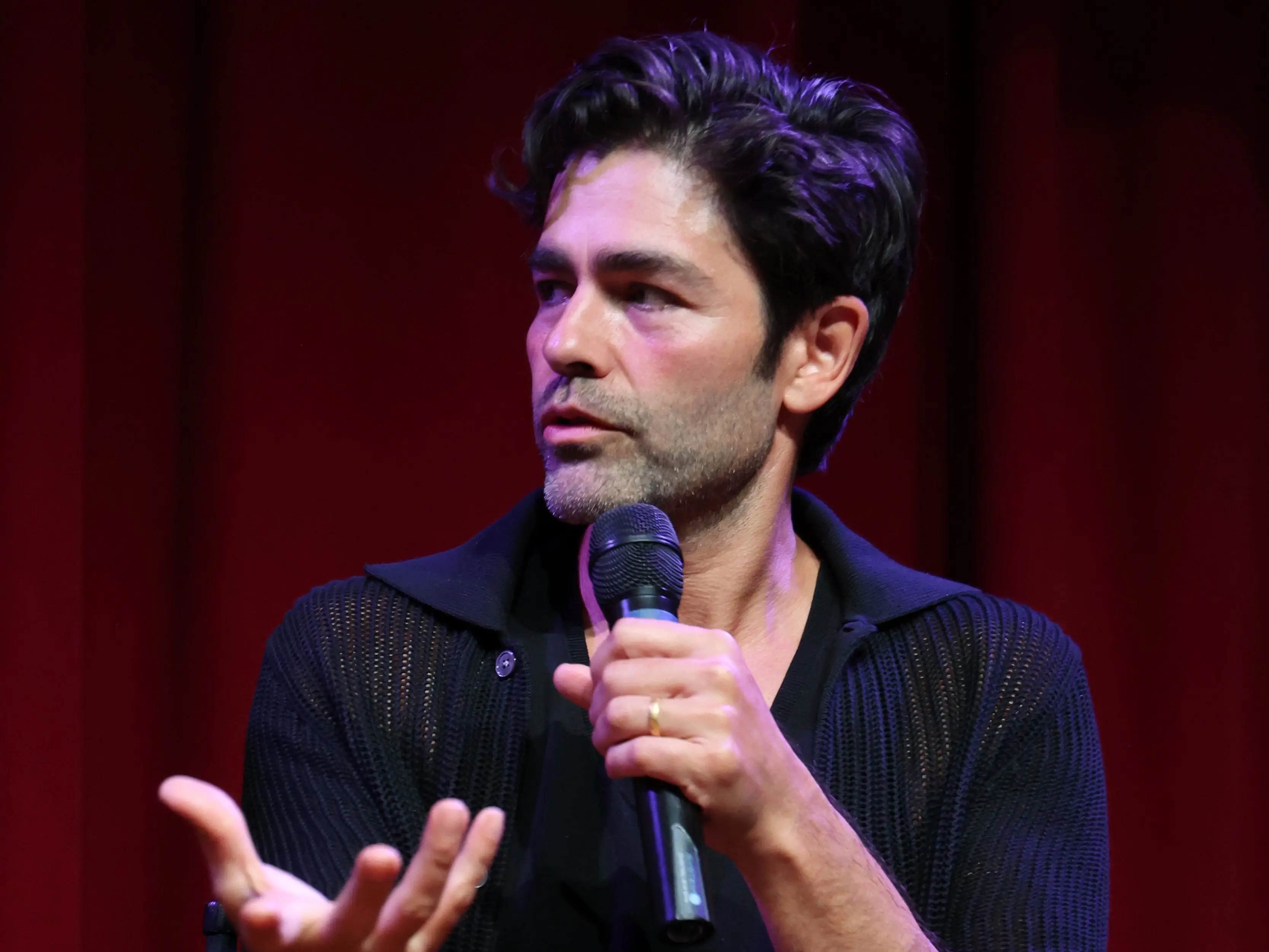 A man holds a microphone on stage, and red drapes flow in the background.