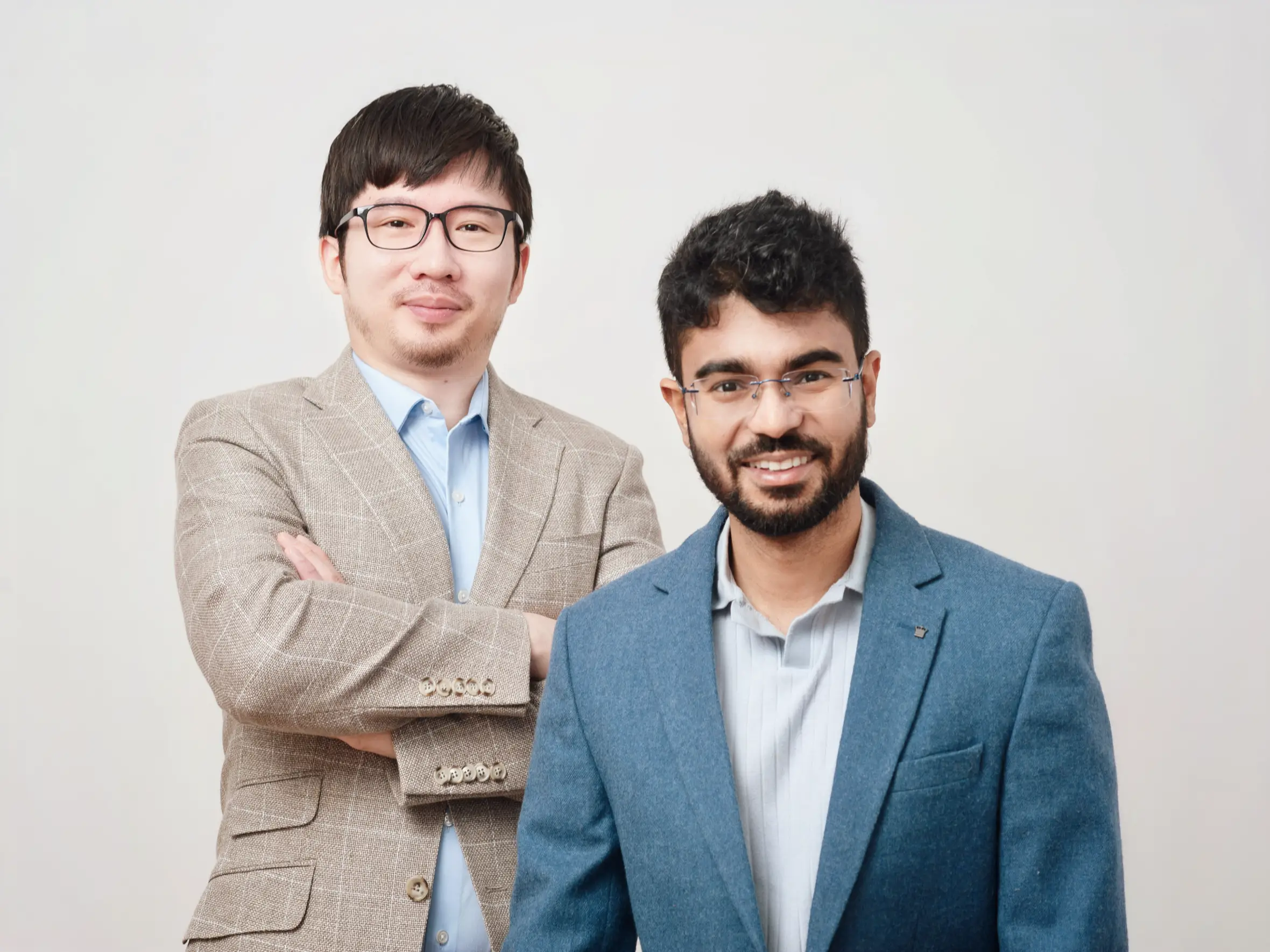 Two men wearing glasses and business attire stand side by side, smiling against a plain background.
