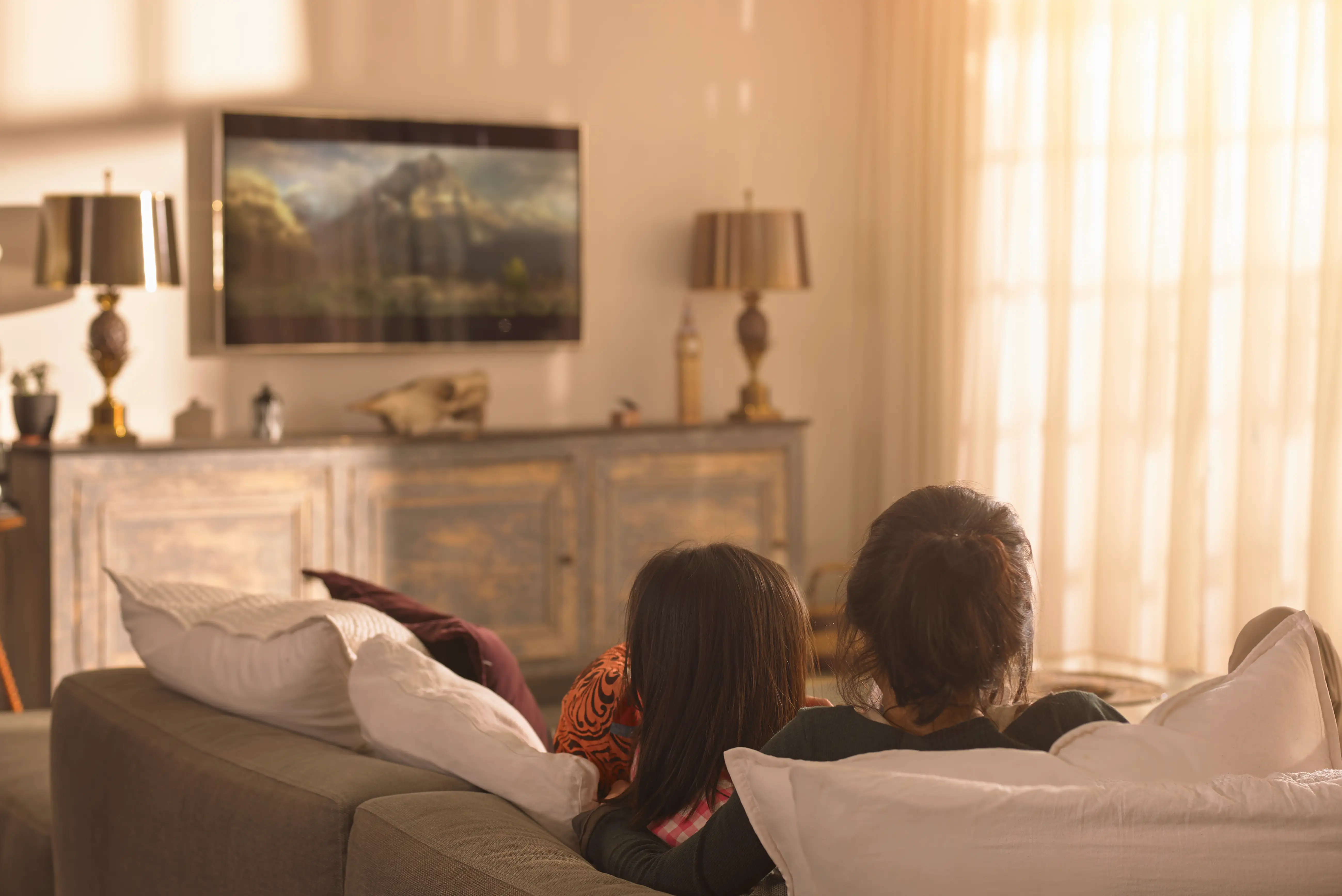 A mother and daughter sit on a couch watching TV together.