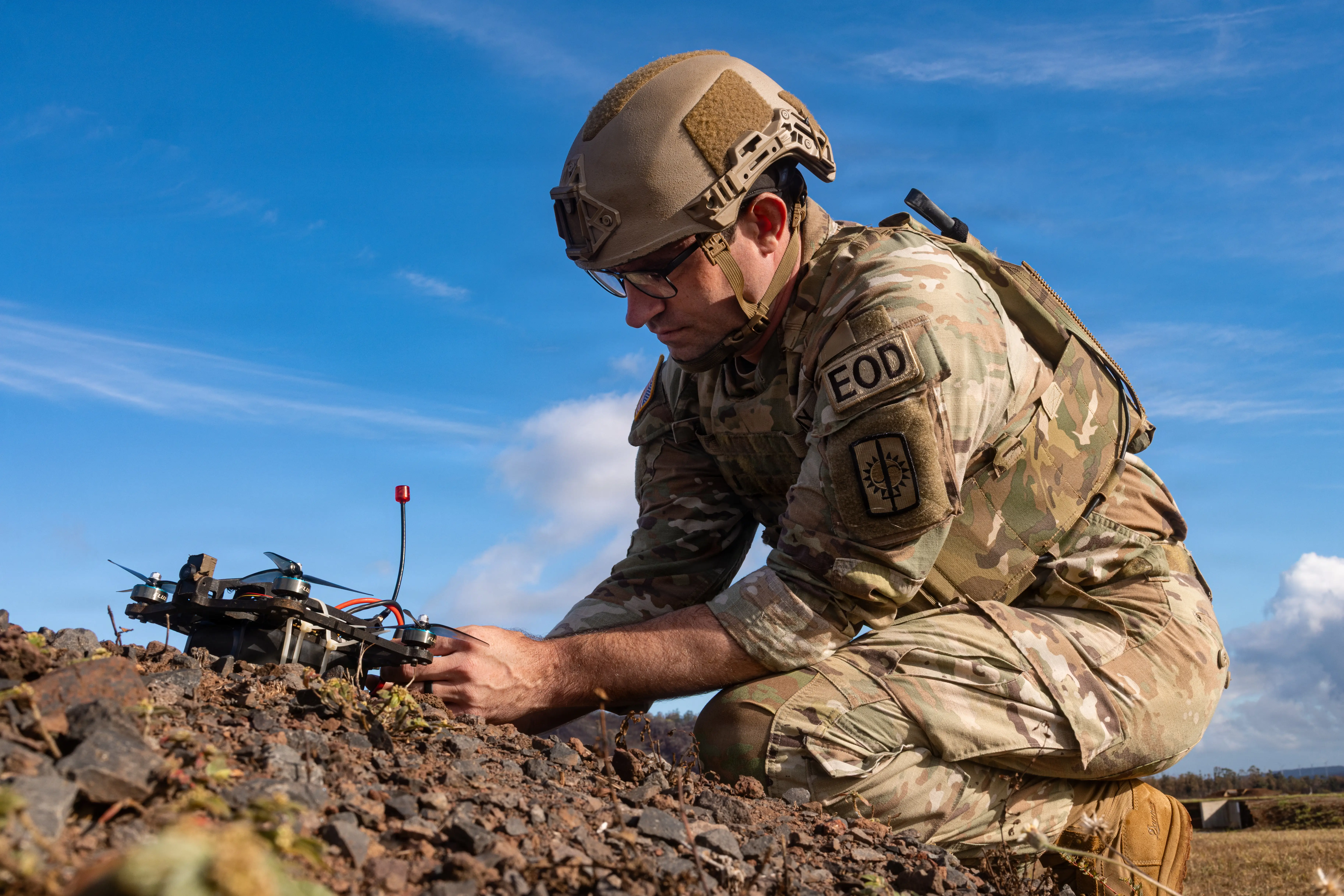 A soldier wearing camouflage crouches and works on a small drone with a blue cloudy sky in the background.