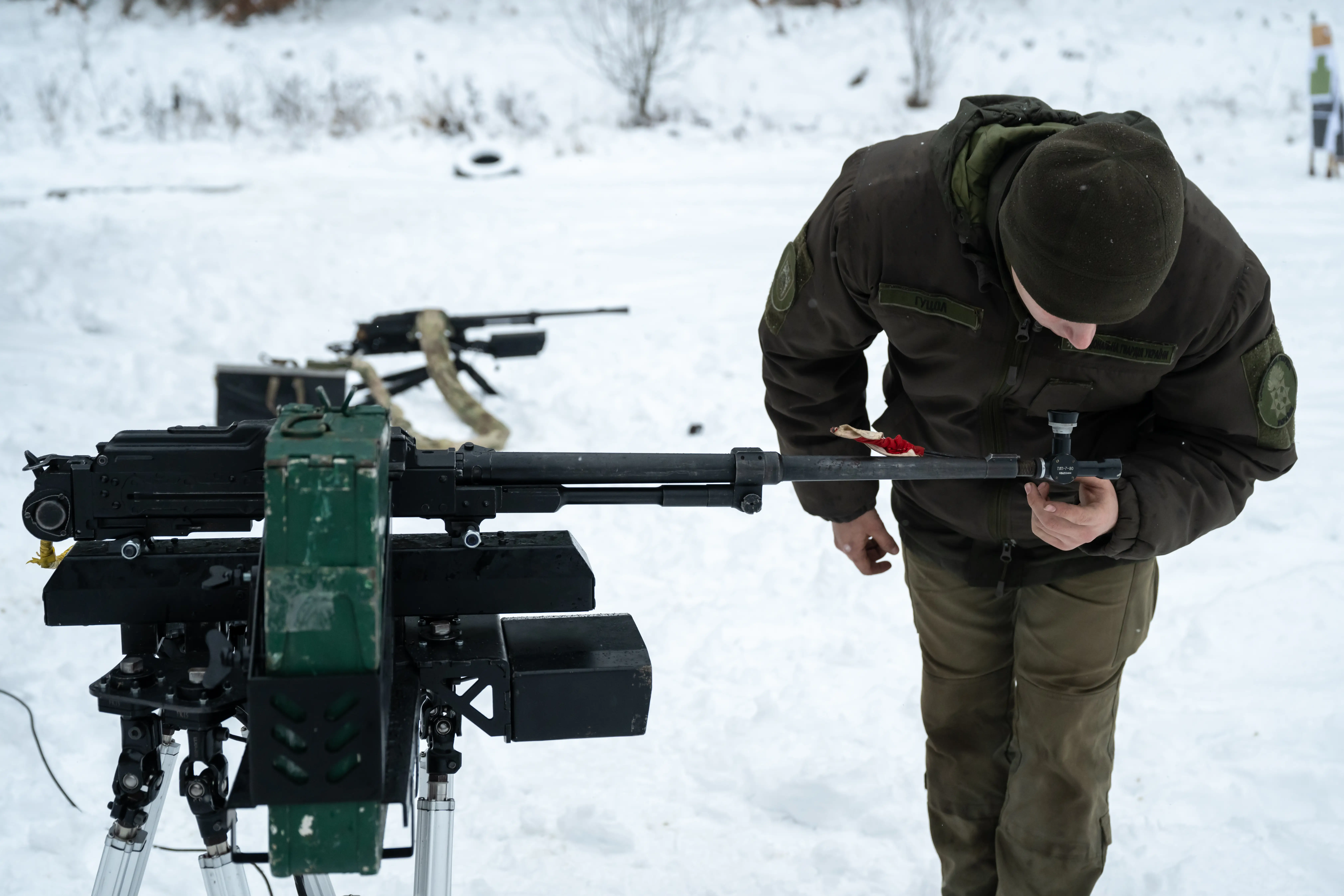 A man in khaki bends over and looks at a robotic system in the snow