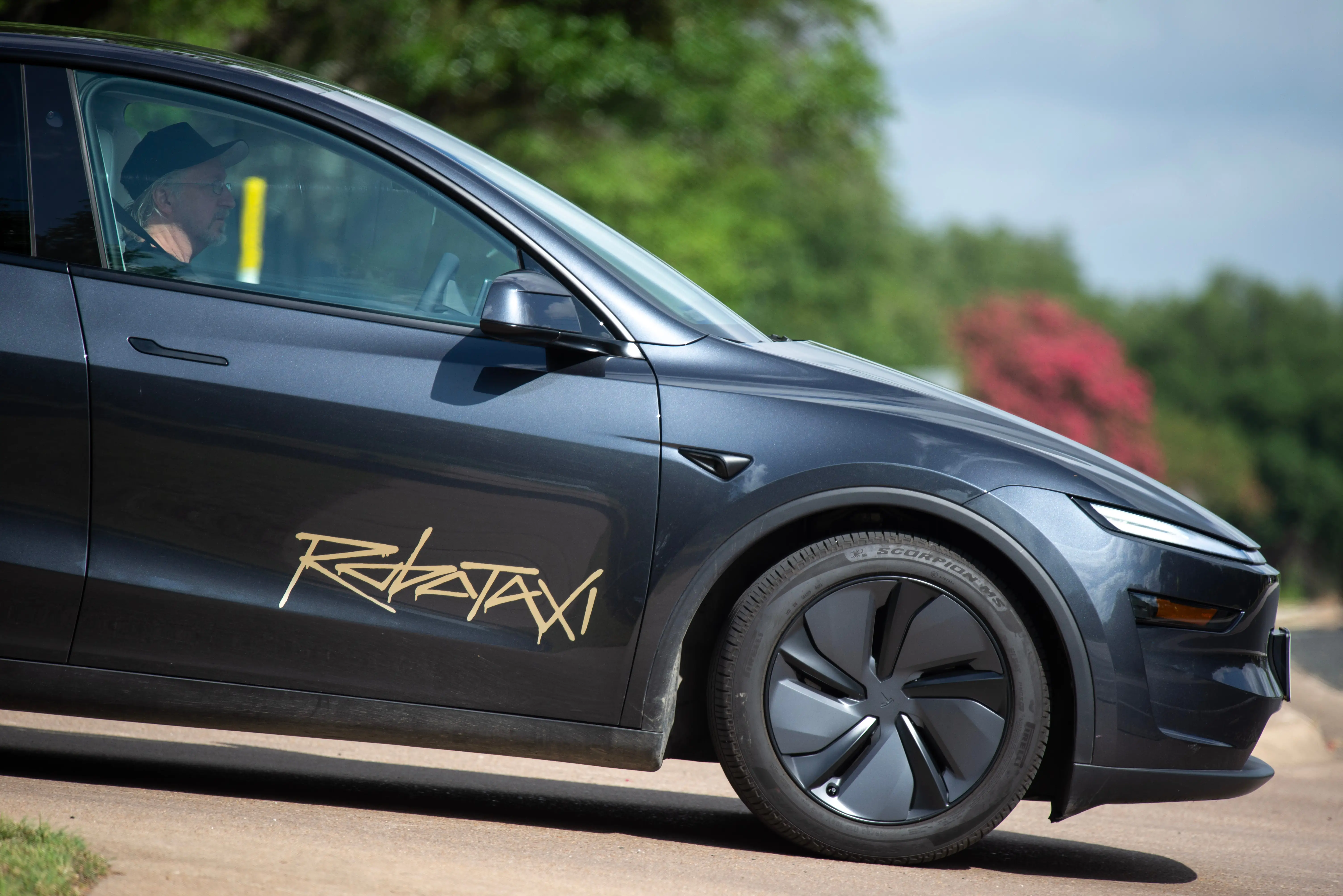 A gray Tesla Model Y robotaxi approaches an intersection with a person sitting in the front seat.
