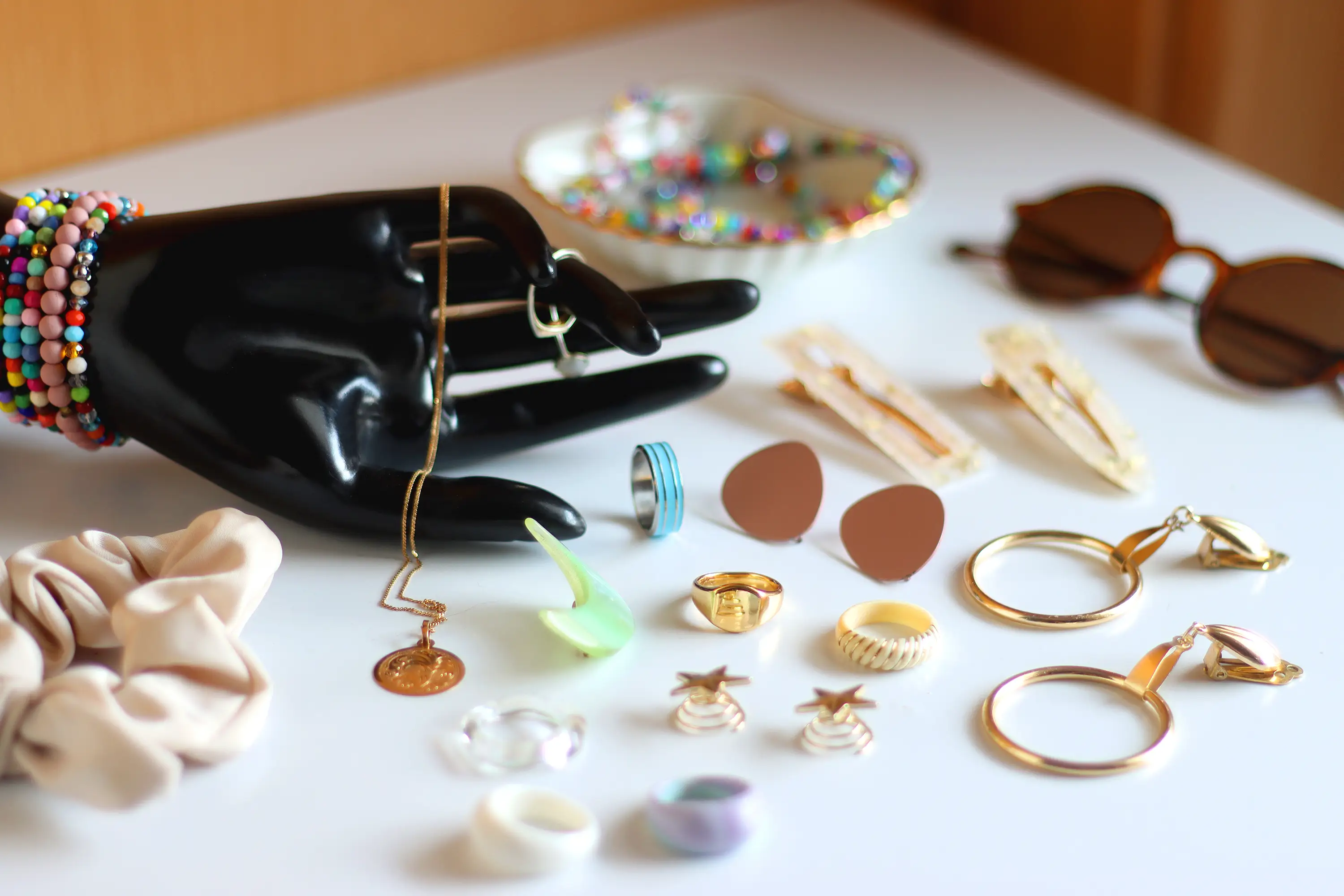 Hand figurine, sunglasses, gold jewelry, colorful bead jewelry, statement rings and various hair accessories on the white table.